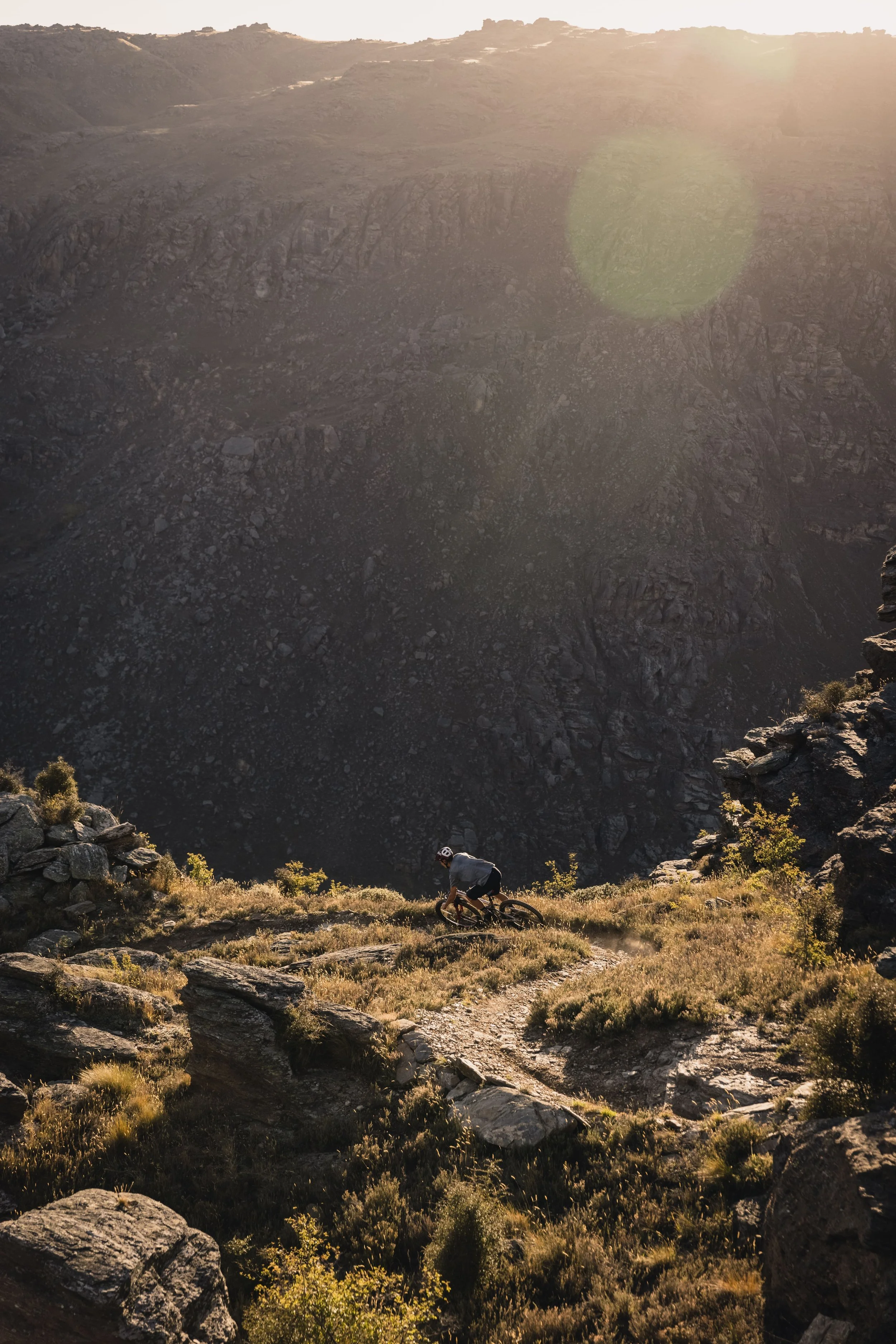 A mountain biker riding along a dirt trail on a rocky hillside with a large mountain or volcanic crater in the background, and the sun shining brightly from the upper right corner, creating a lens flare.