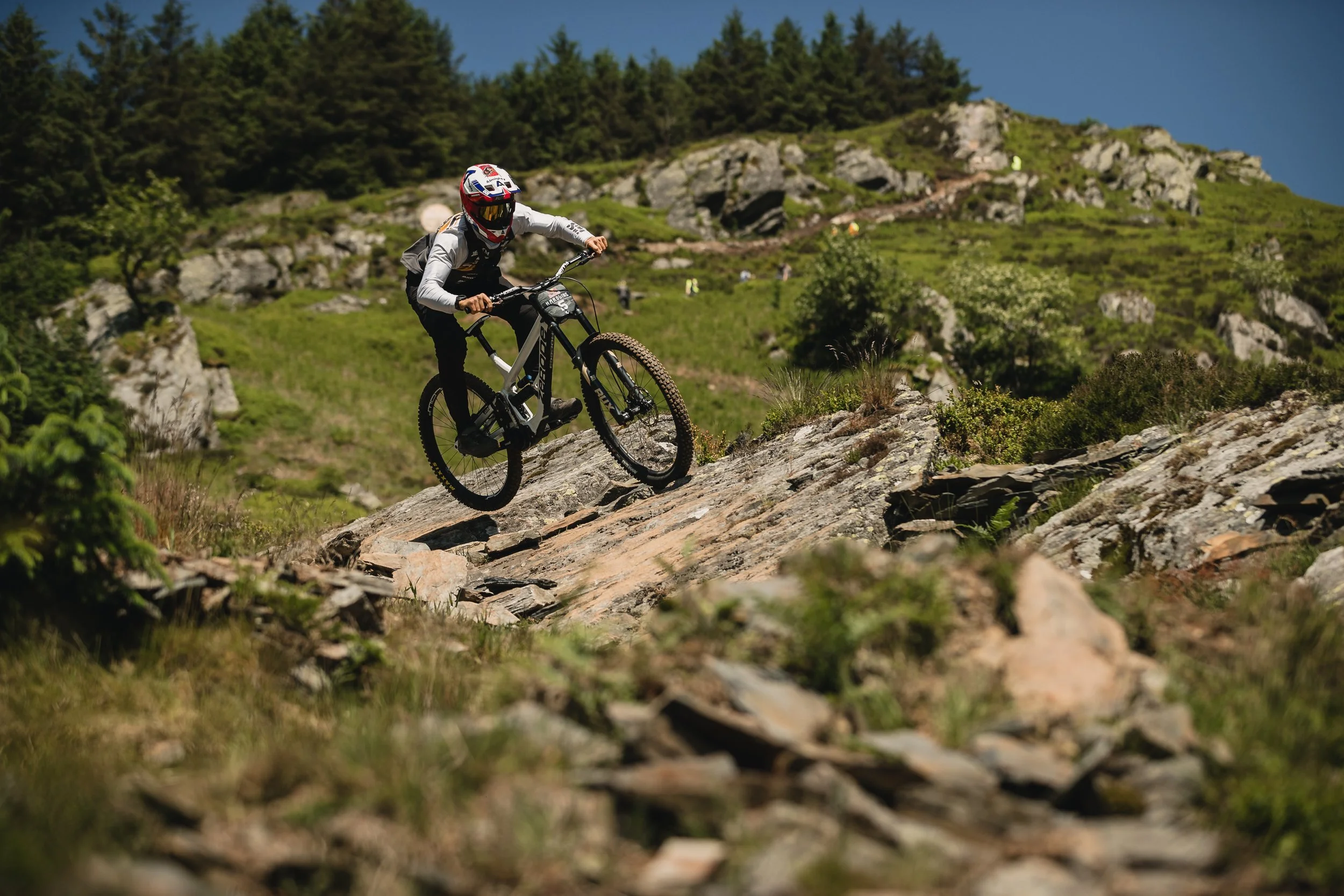 A person mountain biking on a rocky trail through a green hillside with trees and a clear blue sky.
