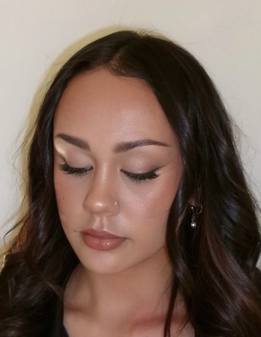 Close-up of a woman with dark wavy hair, closed eyes, natural makeup, and a small nose piercing, against a plain background.