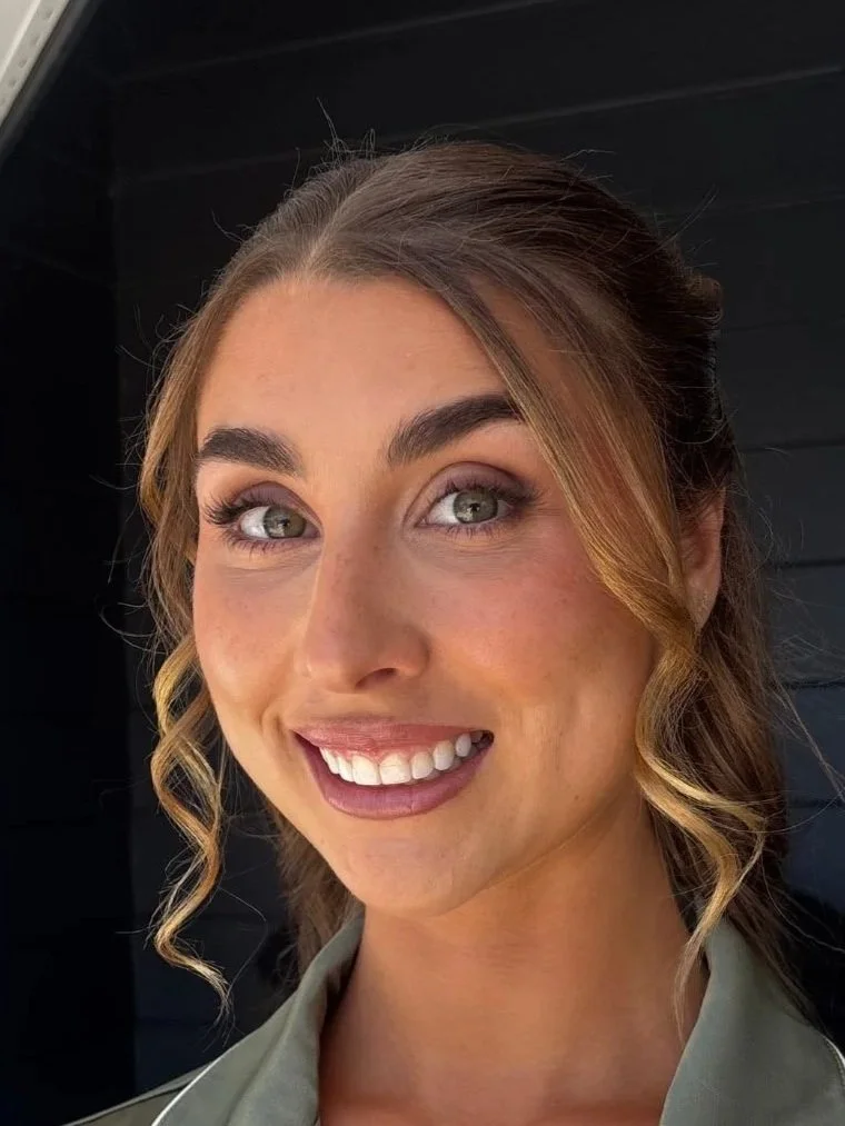 Close-up of a woman with wavy light brown hair, makeup, and a bright smile, standing in front of a dark background.