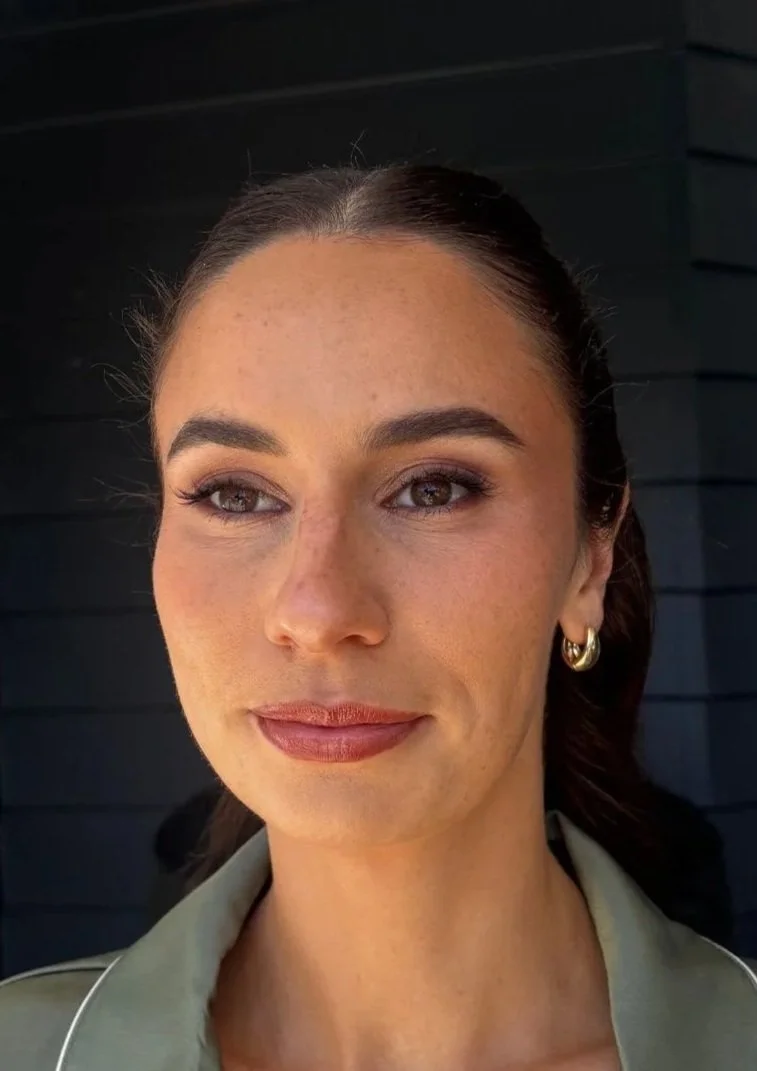 Close-up portrait of a woman with brown hair tied back, wearing gold hoop earrings and a light-colored shirt, standing outdoors against a dark background.