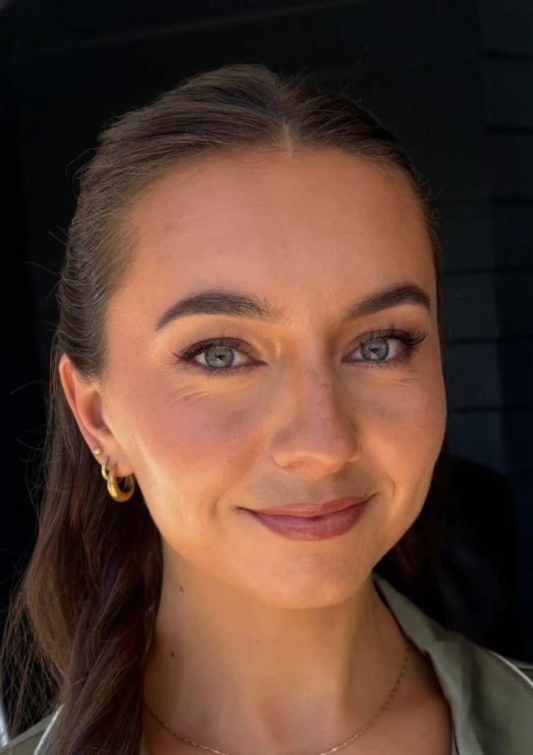 A woman with light skin, long brown hair, and blue eyes, smiling softly, wearing gold earrings and a gold necklace, with natural makeup, shot in close-up against a dark background.