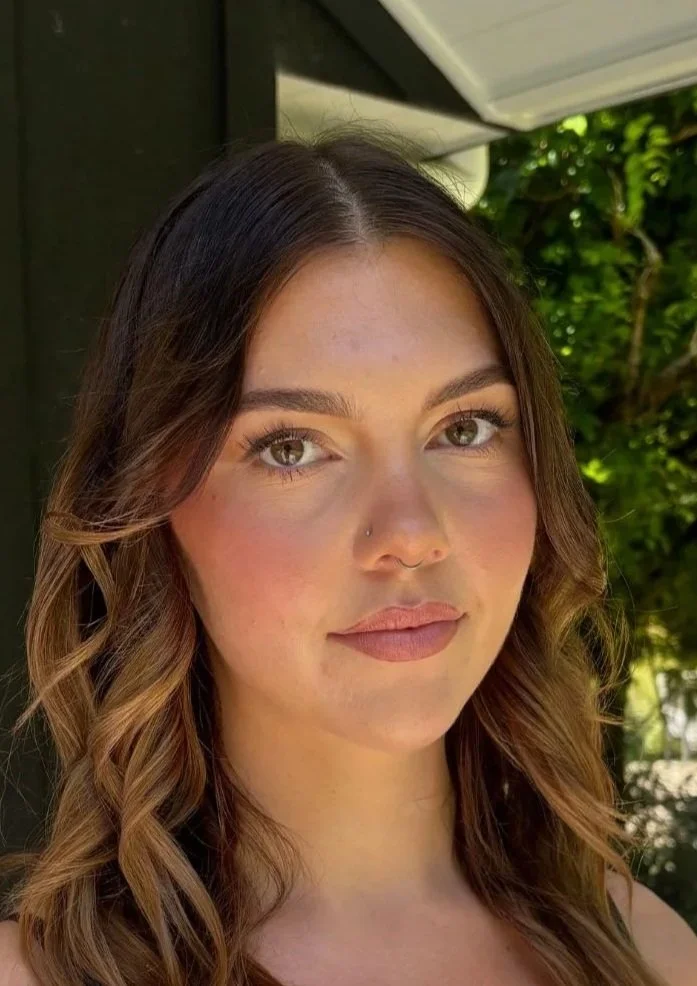 Close-up of a young woman with brown hair, hazel eyes, and a nose piercing, standing outdoors near greenery.