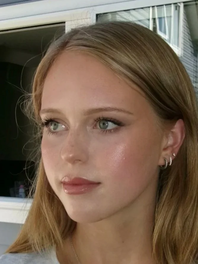 Close-up of a young woman with long, red hair and light makeup, looking slightly to the side, posing outdoors near a building with large windows.