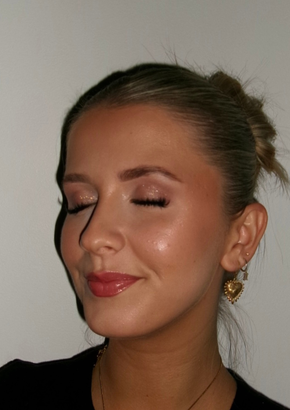 A woman with her eyes closed, smiling, with makeup, earrings, and a bun hairstyle, against a plain background.