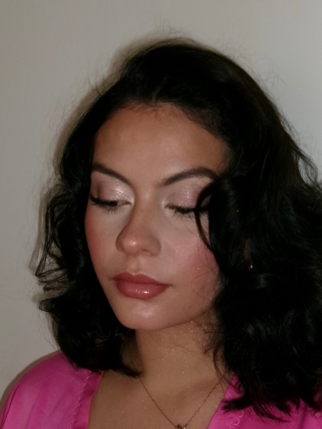 A woman with black wavy hair and light makeup, wearing a pink top, with her eyes closed and a neutral expression on her face.