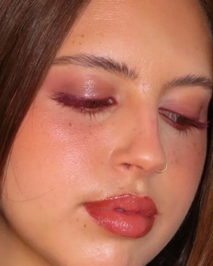 Close-up of a woman's face showing her makeup, including pink eyeshadow, glossy lips, and freckles.