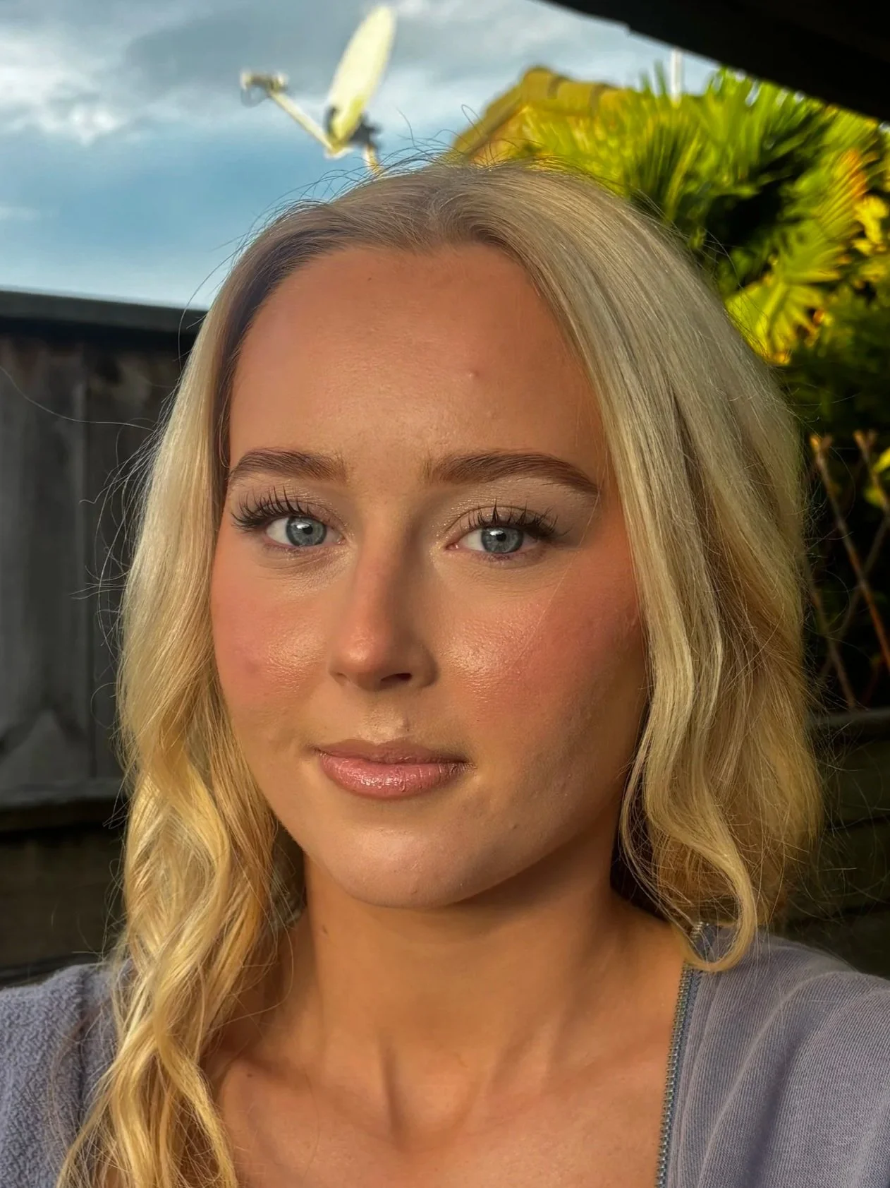 A close-up of a young woman with long blonde wavy hair, blue eyes, and light makeup, standing outdoors with a wooden fence, green leaves, and a partly cloudy sky in the background.