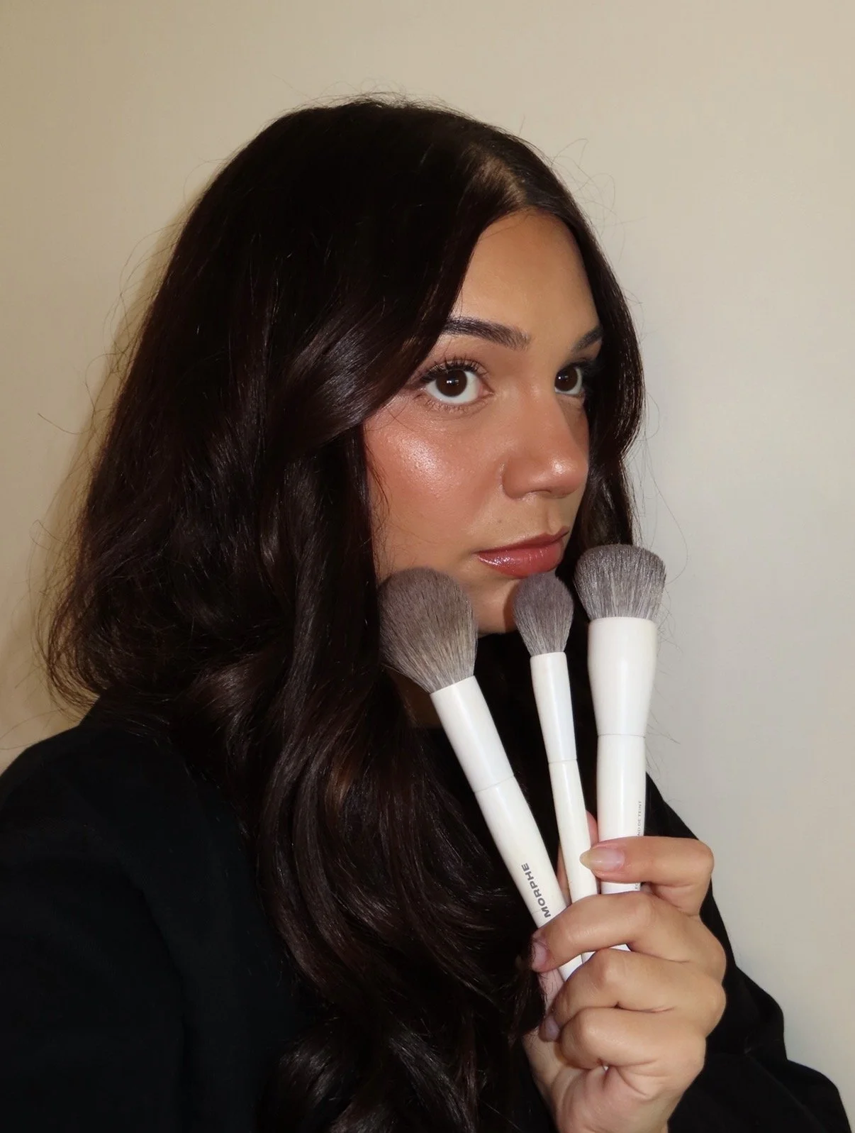 Woman with long dark hair holding three makeup brushes close to her face, standing against a plain wall.