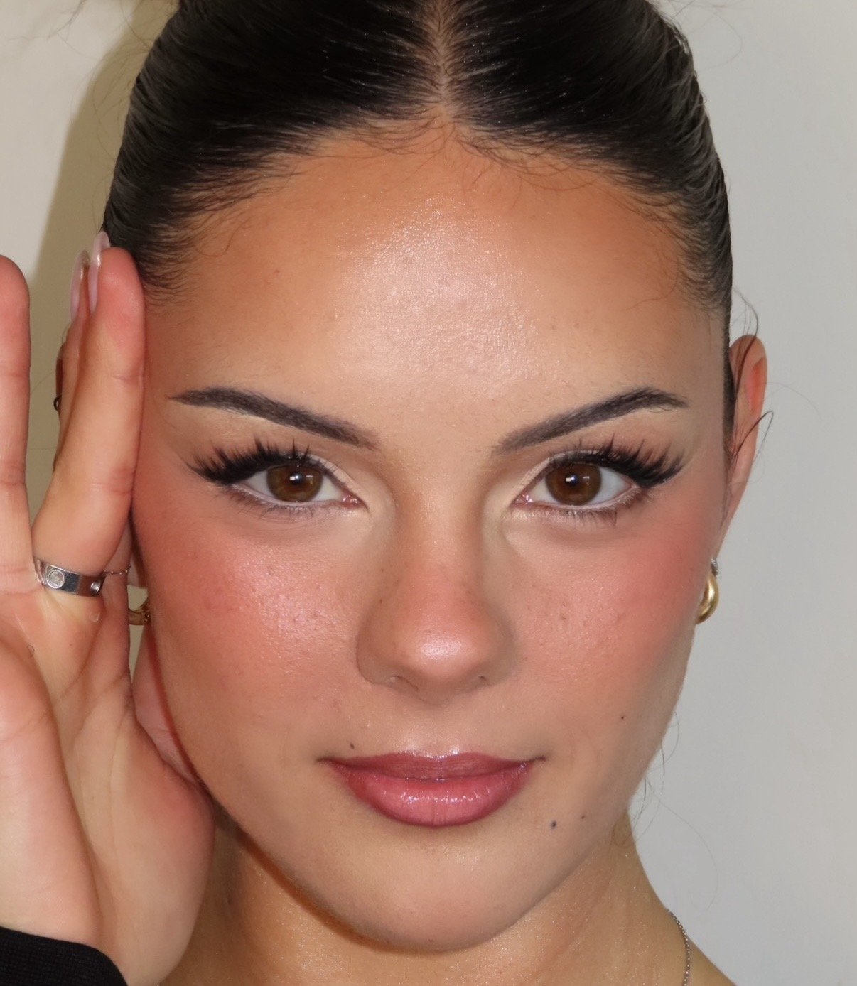 Close-up of a woman's face with styled dark hair, makeup, and brown eyes, holding her head with her left hand, wearing rings and earrings.