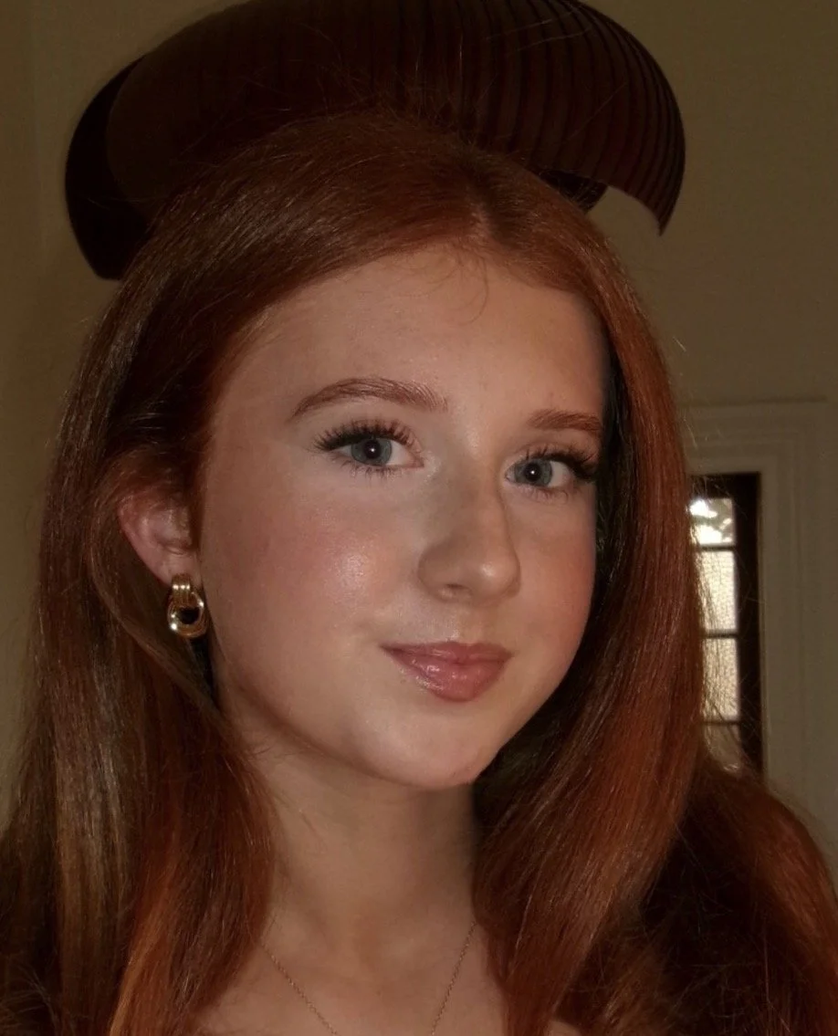Young woman with long red hair, wearing gold hoop earrings, in front of a window with a shade, illuminated by warm indoor lighting.
