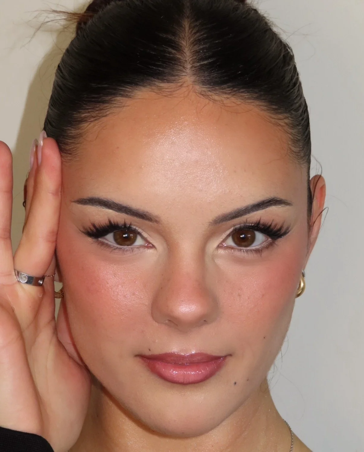 Close-up portrait of a woman with dark hair tied back, brown eyes, makeup, and jewelry, holding her hand near her face against a plain background.