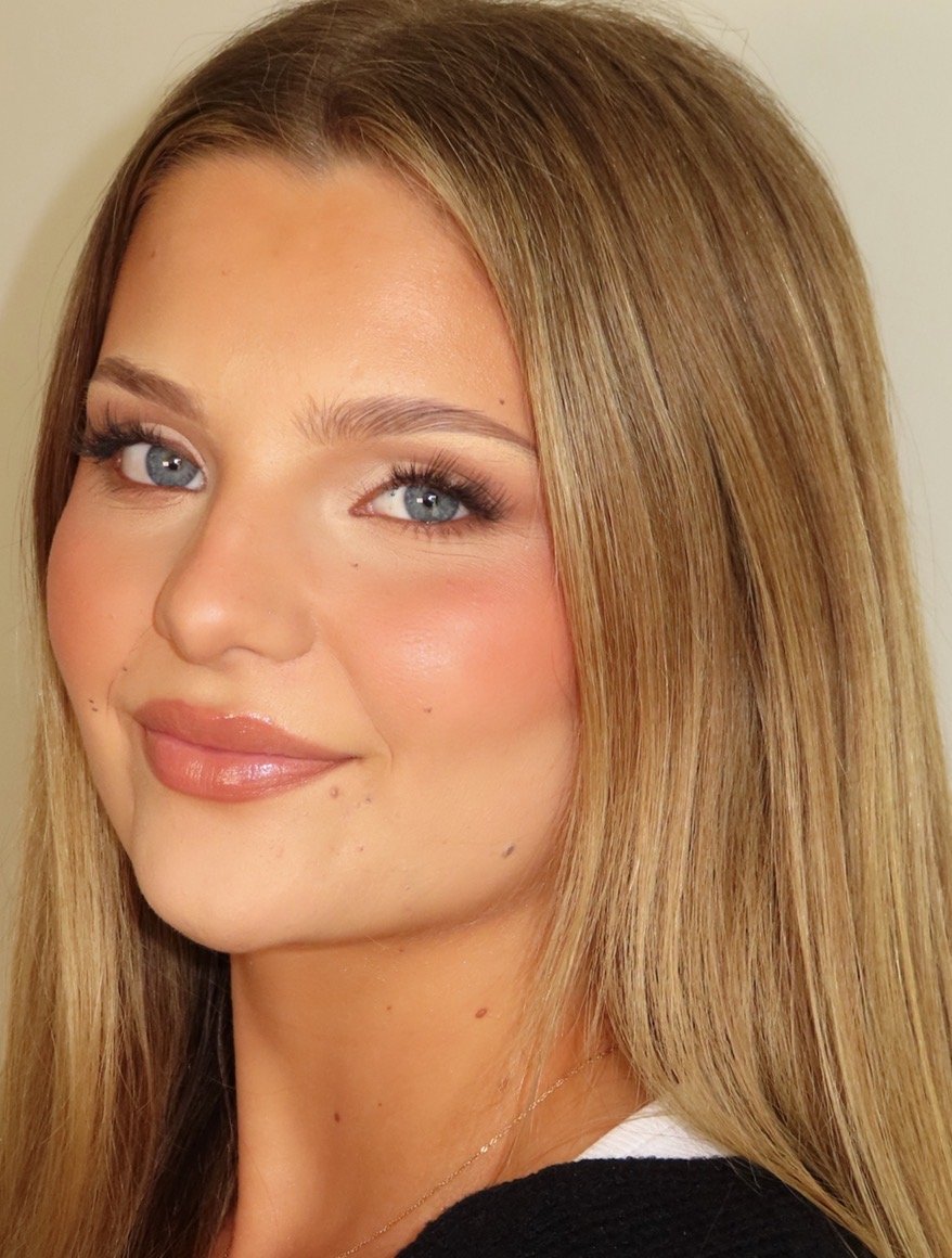 Close-up of a young woman with long blonde hair, blue eyes, and makeup including foundation, blush, and lipstick, smiling softly.
