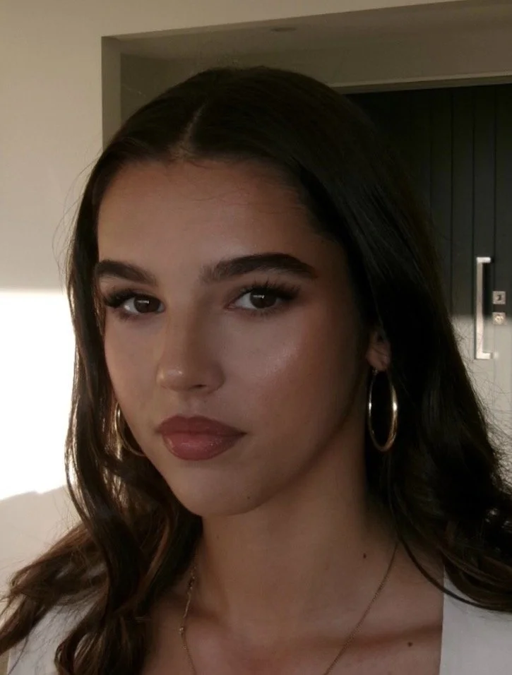 Close-up of a young woman with dark brown hair and gold hoop earrings, looking at the camera indoors.