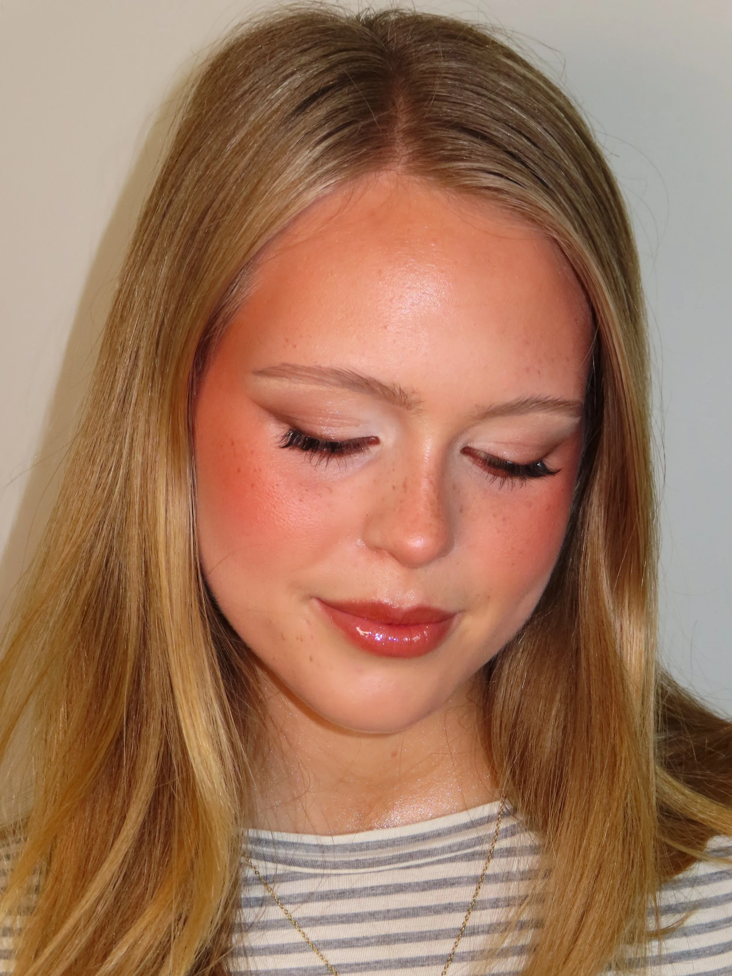 Close-up of a woman with long blonde hair, wearing a striped shirt and a gold necklace, smiling with closed eyes.
