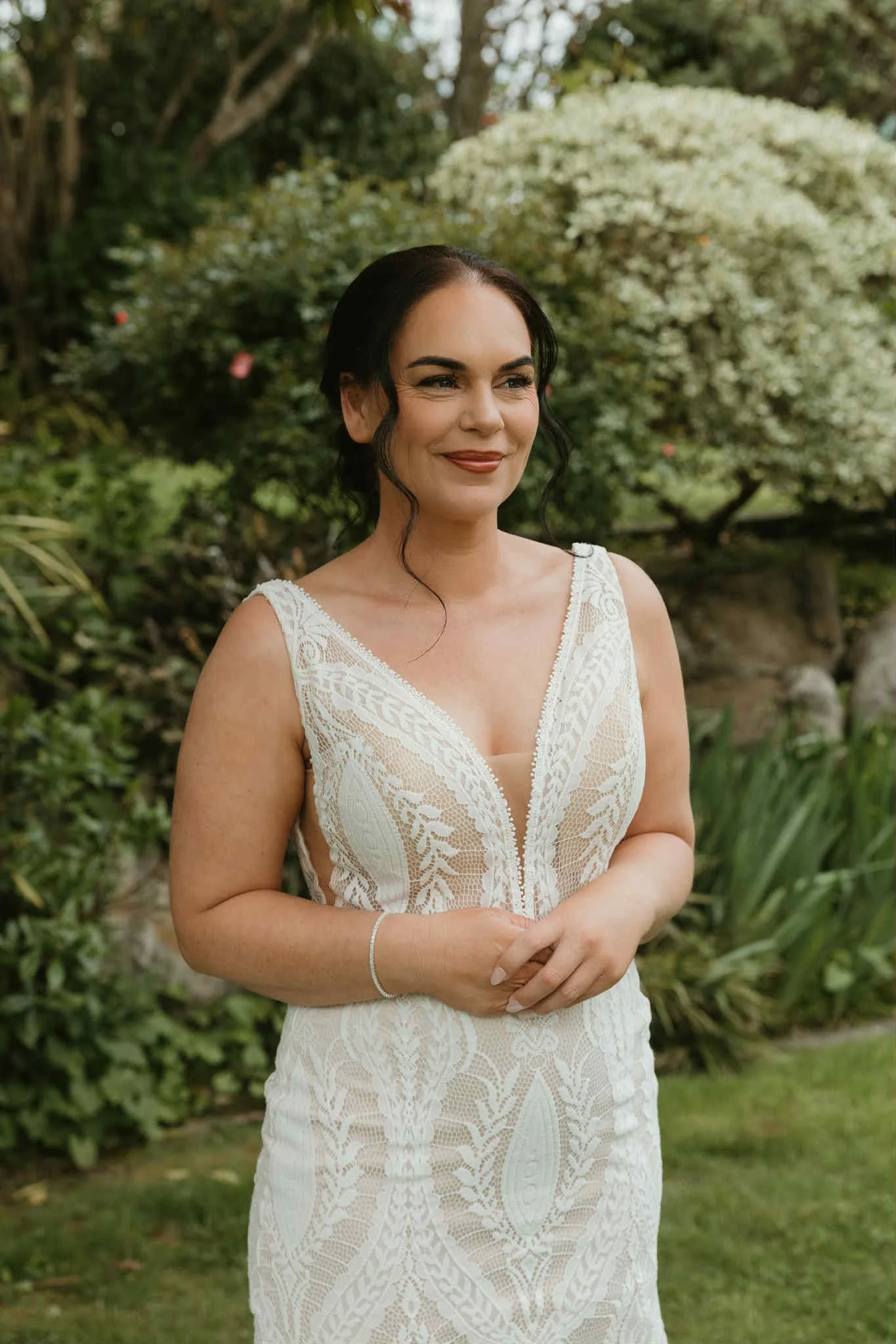 A woman with dark hair in a low bun, wearing a sleeveless white lace dress, standing outdoors in a garden with trees and white flowers in the background.
