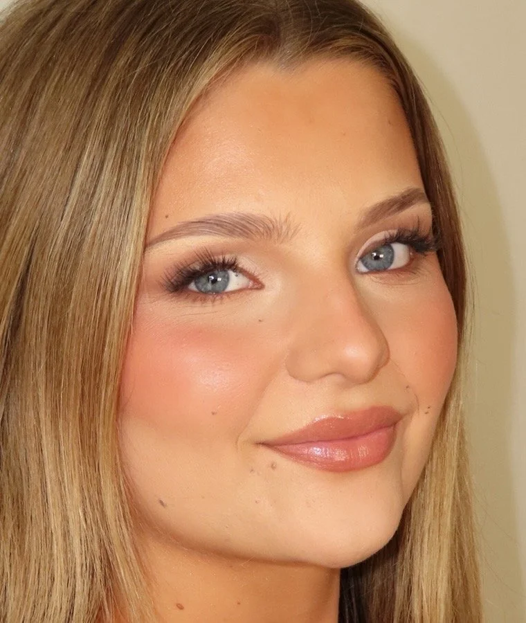 Close-up of a young woman with blonde hair, blue eyes, wearing makeup with pink lipstick and light blush, smiling softly.