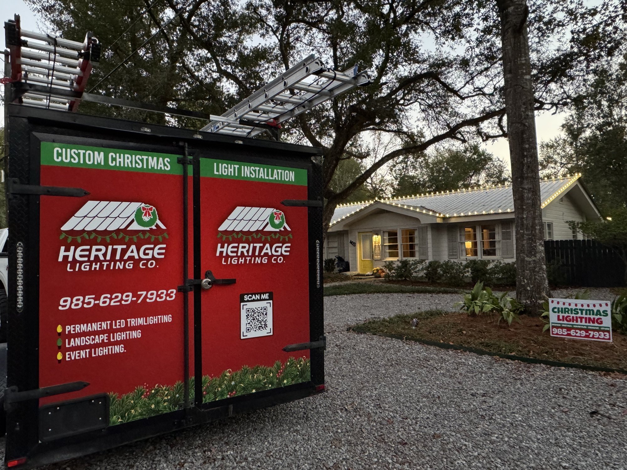 Heritage Lighting Co. trailer with ladders beside a house decorated with warm white Christmas lights