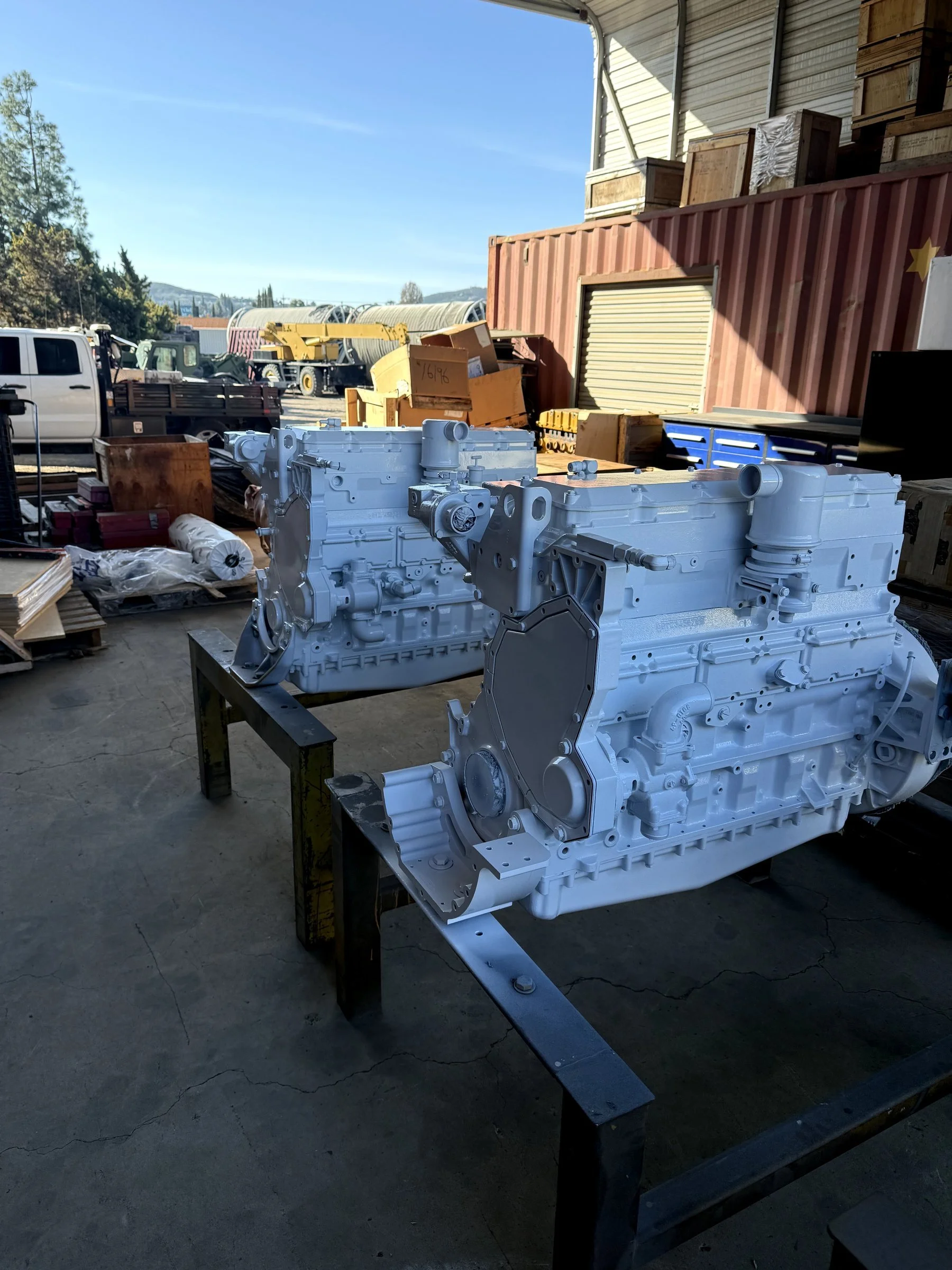 A white mechanical engine on a metal stand inside a workshop or storage area, with wooden boxes and crates, a red shipping container, and vehicles in the background.