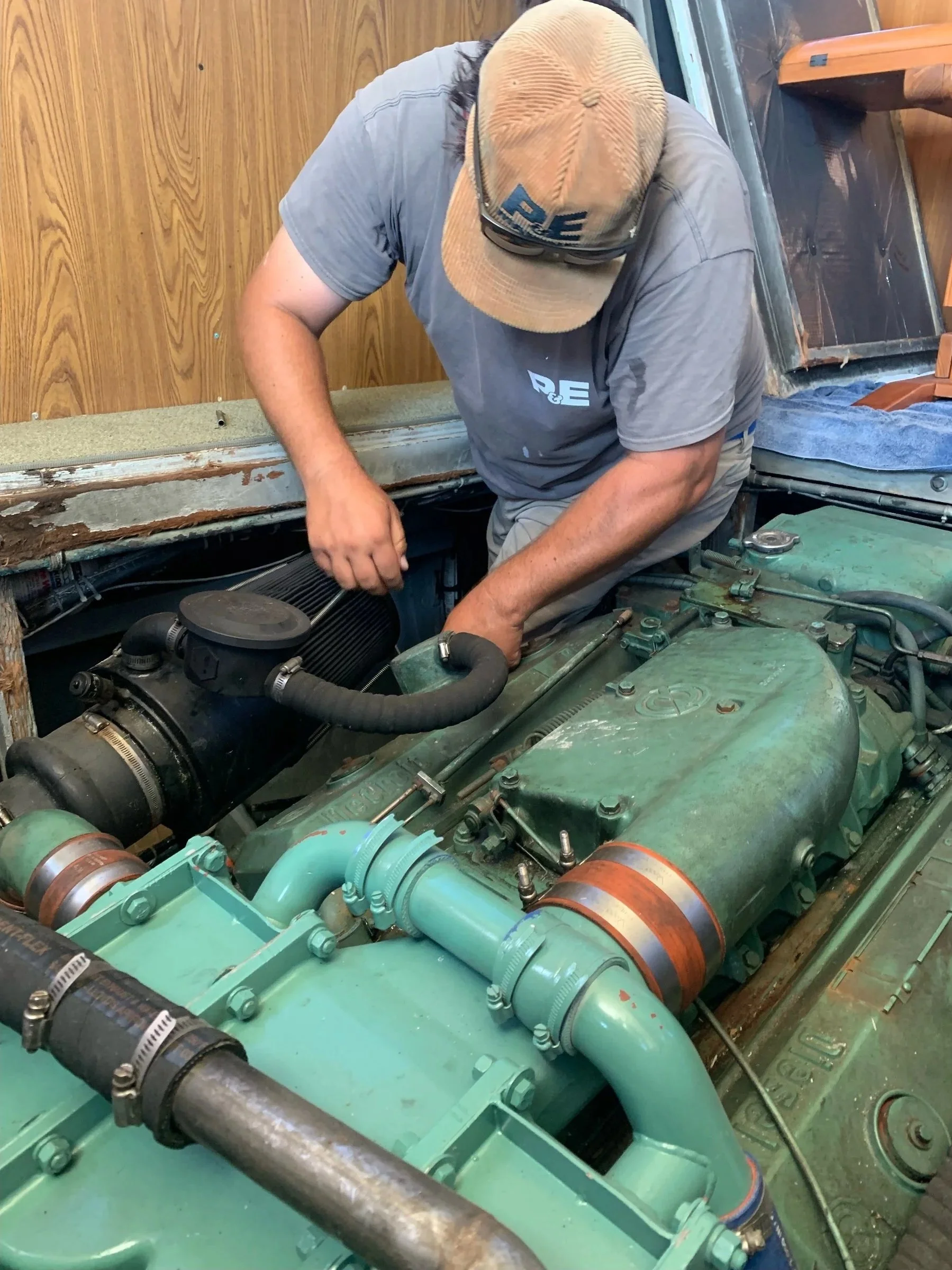 A man wearing a beige cap and gray t-shirt inspecting or working on the engine of a vintage military-style vehicle, possibly an old Jeep, with a green engine block, hoses, and parts.