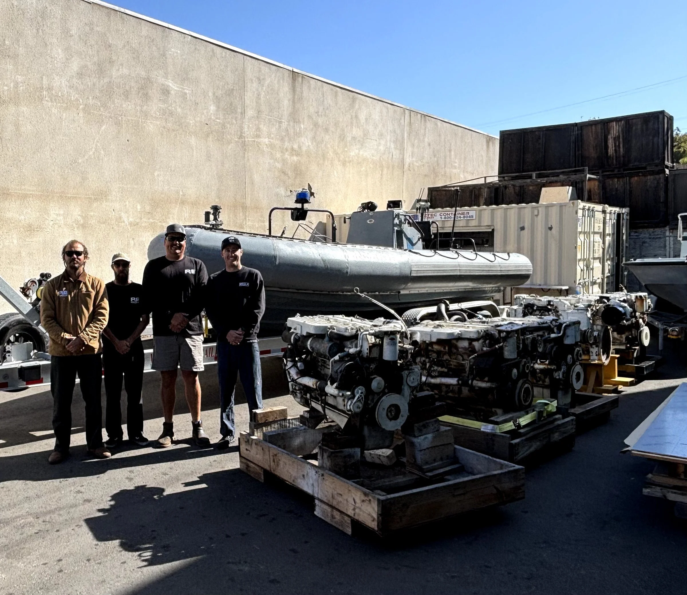 A group of four men standing in front of a submarine on a trailer, with large shipping containers and crates nearby, outside a building with a plain wall and a clear blue sky.