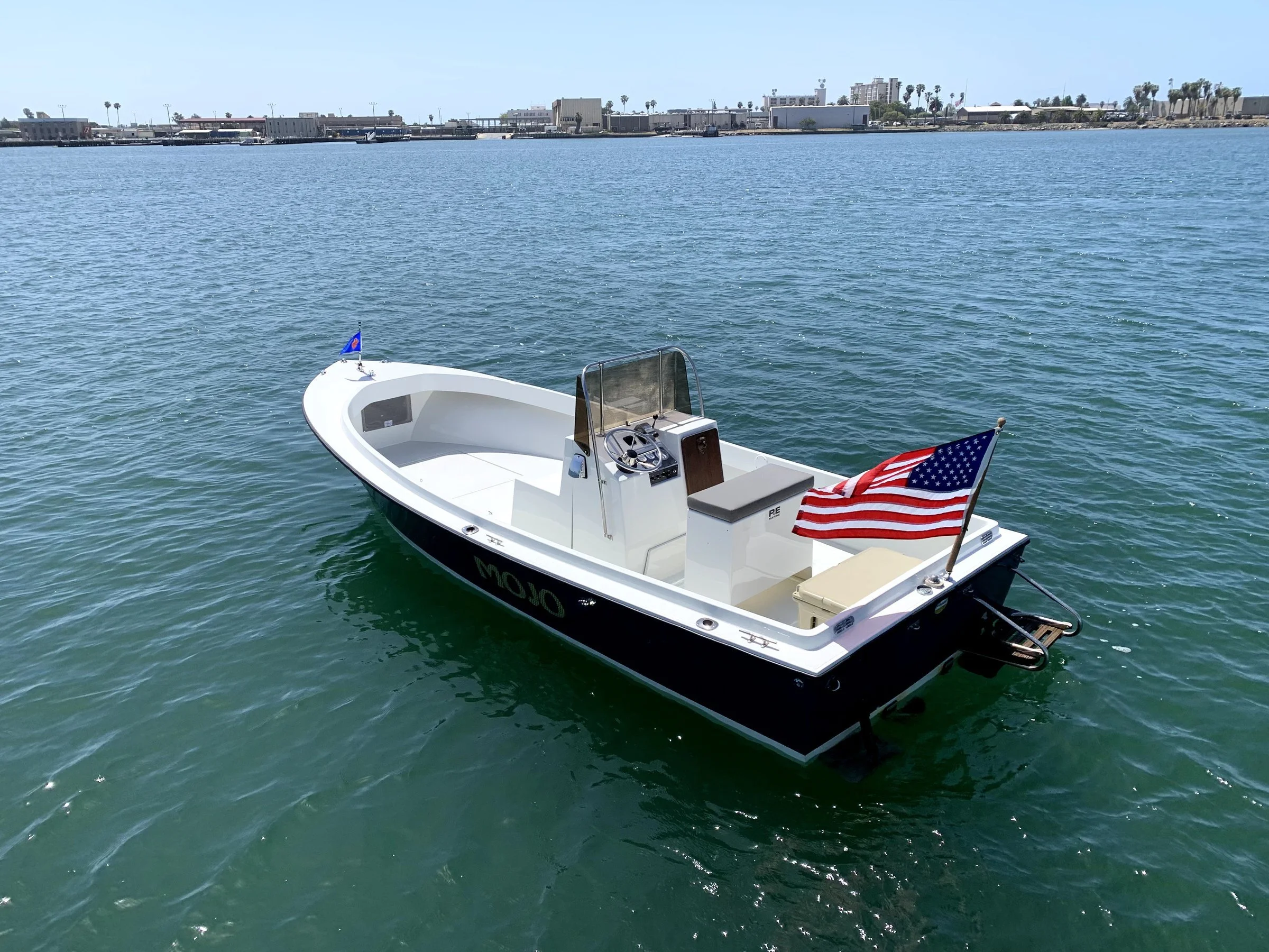 Small white motorboat with an American flag at the back floating on calm water near a shoreline with buildings and palm trees in the background.