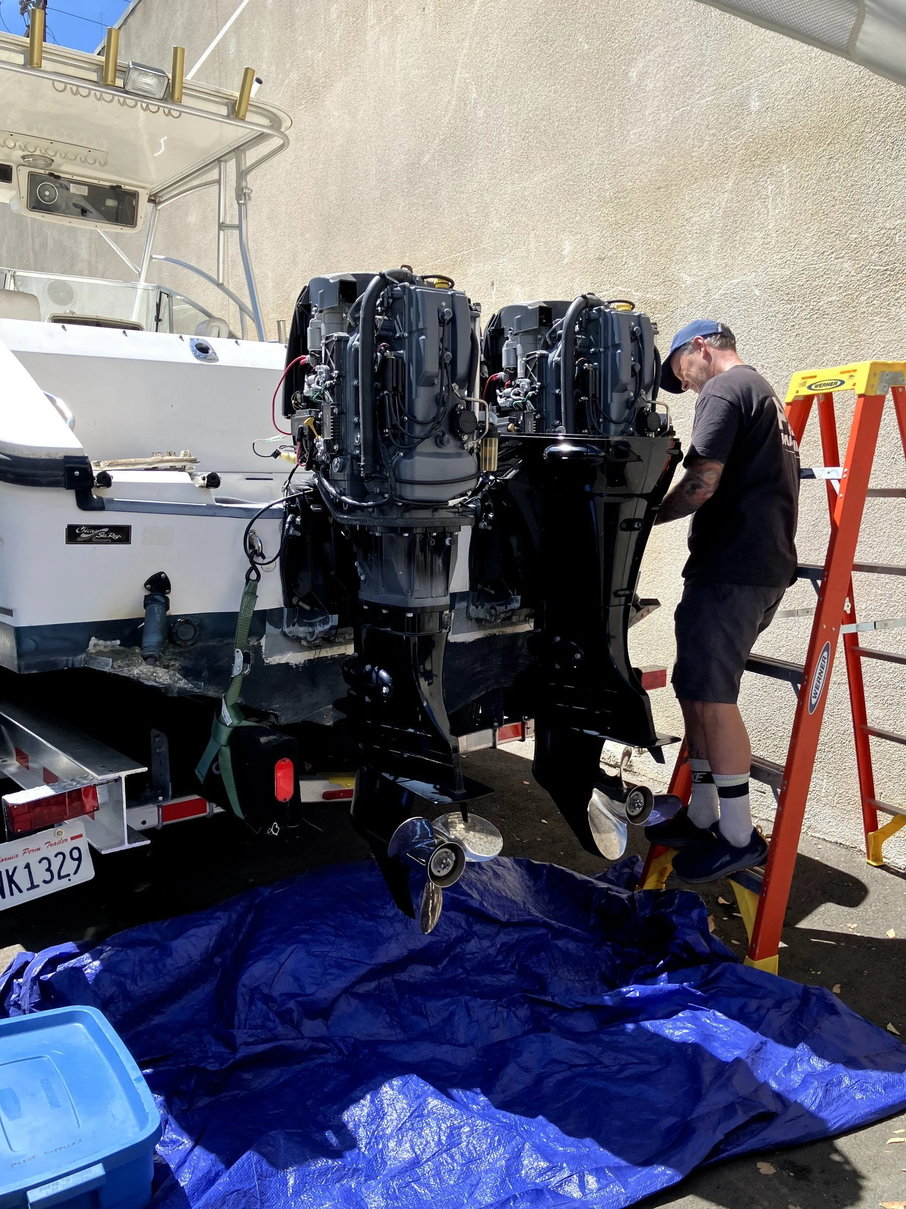 A man working on a boat engine stands on a ladder next to a large boat that is on a trailer, with a blue tarp underneath. The man is wearing a black T-shirt, gray shorts, and a blue cap. The boat has two outboard motors and a white exterior. Various 