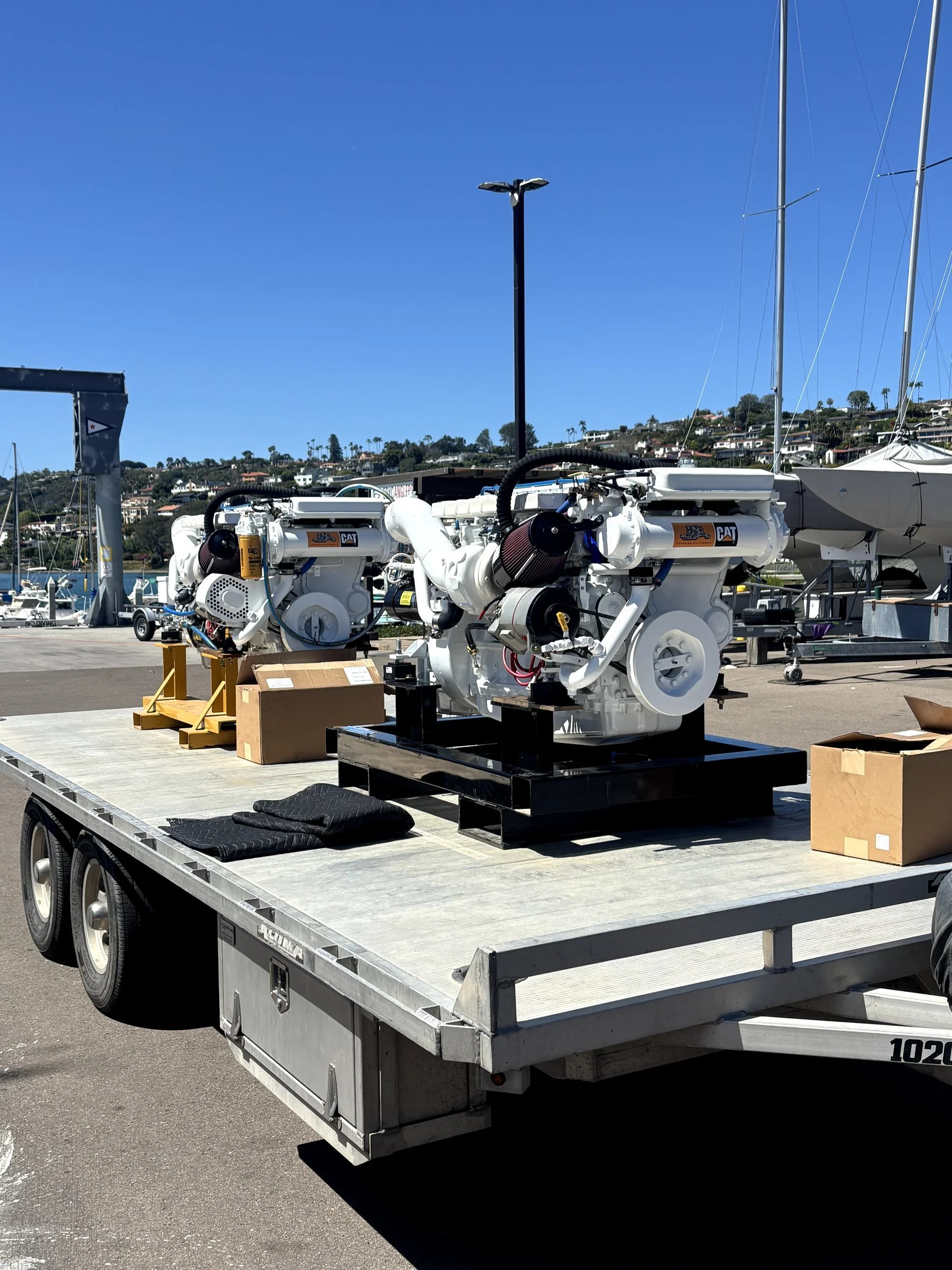A flatbed trailer carrying two large white industrial engines with CAT logos, boxes, and tools, parked outdoors at a marina with sailboats and hillside homes in the background.