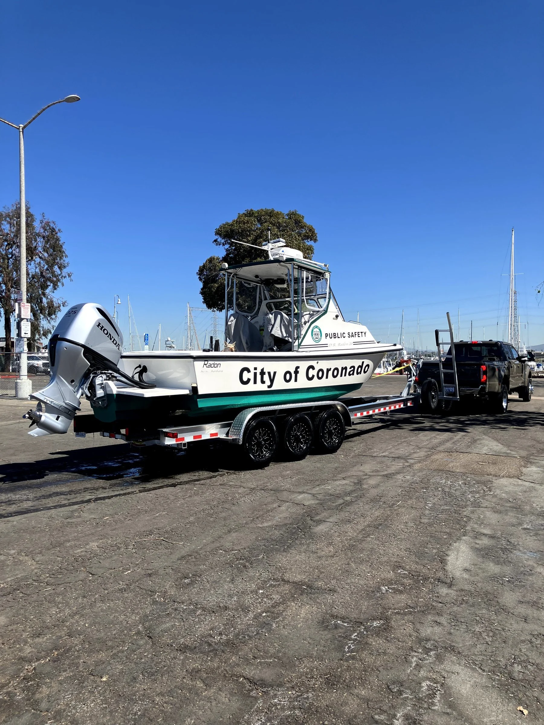 A boat with a Honda outboard motor on a trailer, marked with 'City of Coronado' and 'Public Safety,' parked in a lot with sailboats in the background under a clear blue sky.