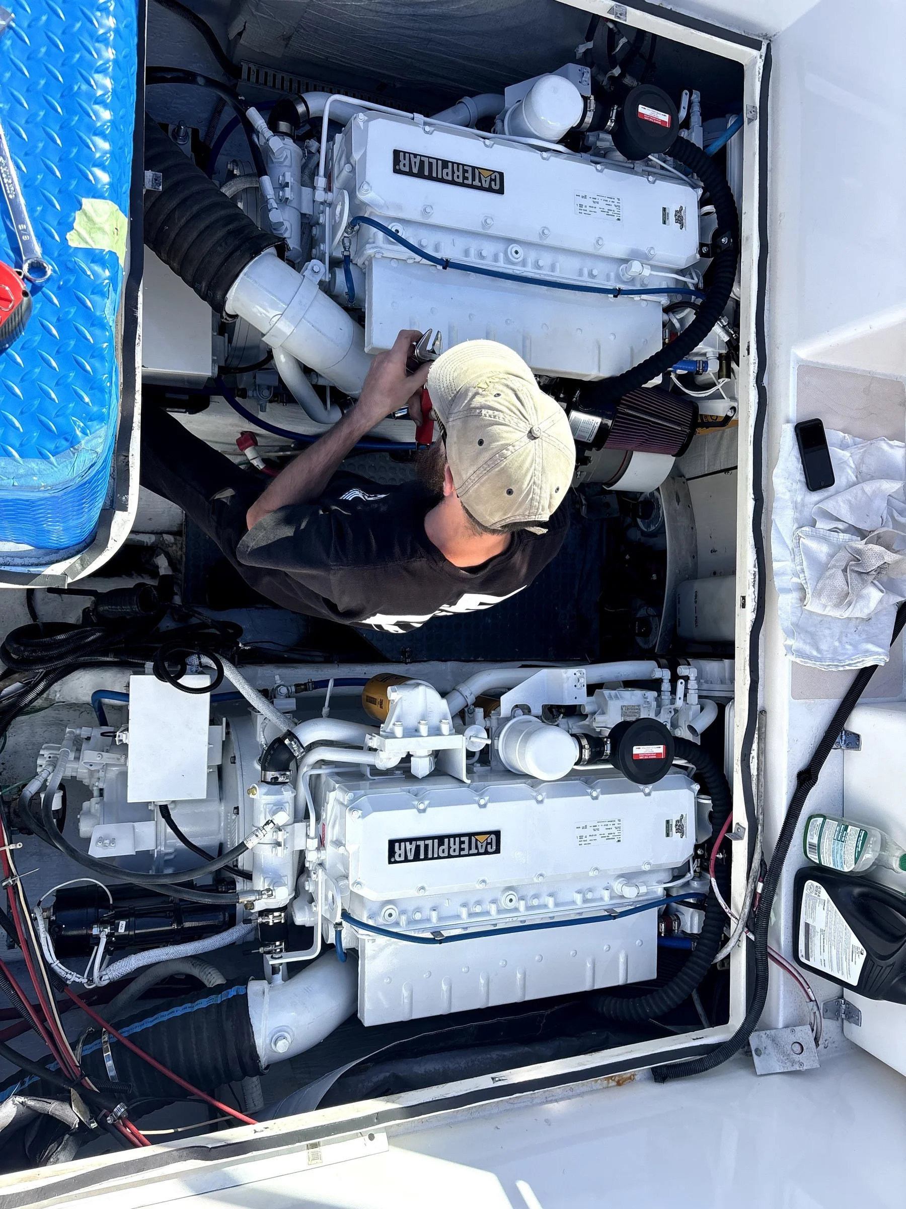 A person working underneath a Caterpillar engine, possibly on a boat or vehicle, with various parts and tools visible.