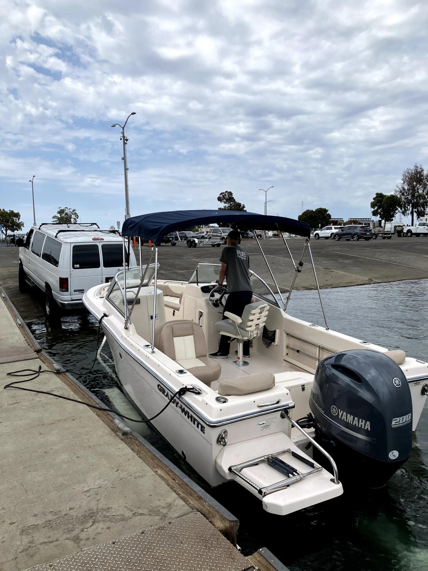 A white boat with a blue canopy docked at a marina with a man standing inside, surrounded by parked cars and lampposts, under a cloudy sky.