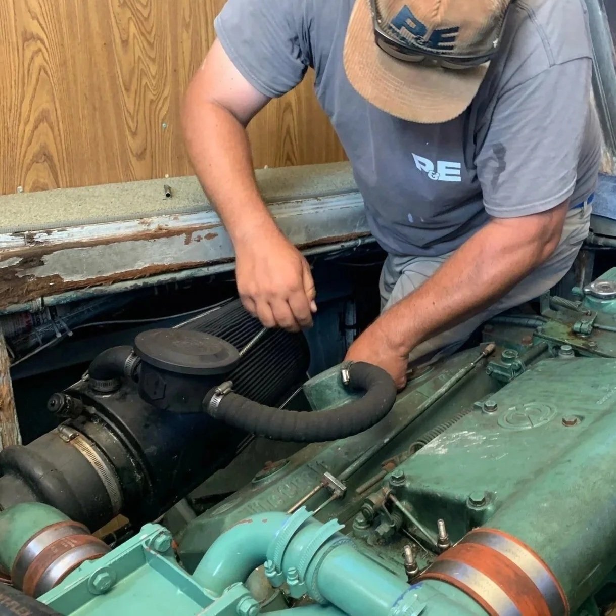 A man wearing a cap and gray t-shirt working on a vehicle engine in a garage, with tools and engine components visible.