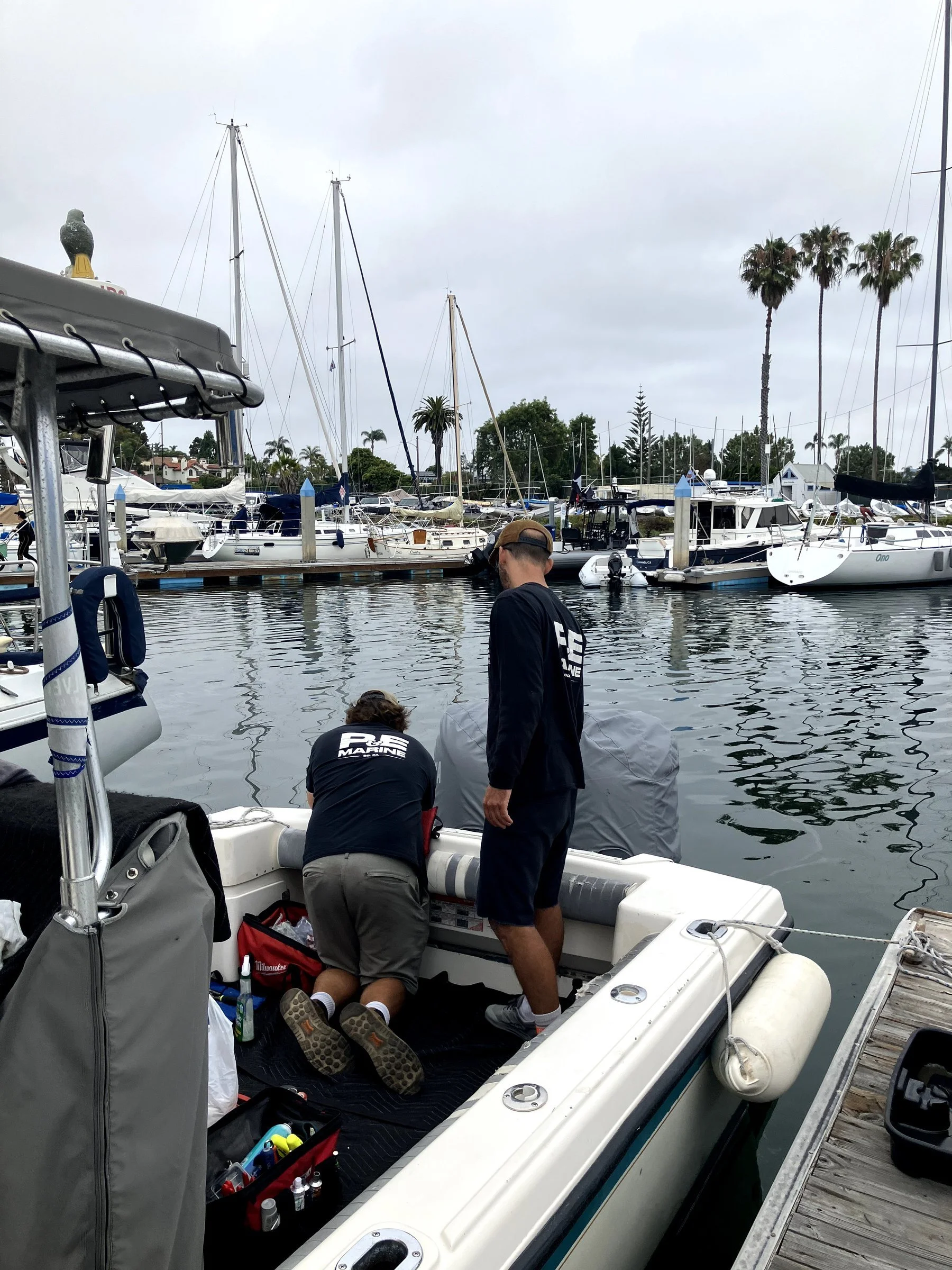 Two men preparing a boat for launch at a marina, with several boats docked in the water and palm trees in the background under an overcast sky.