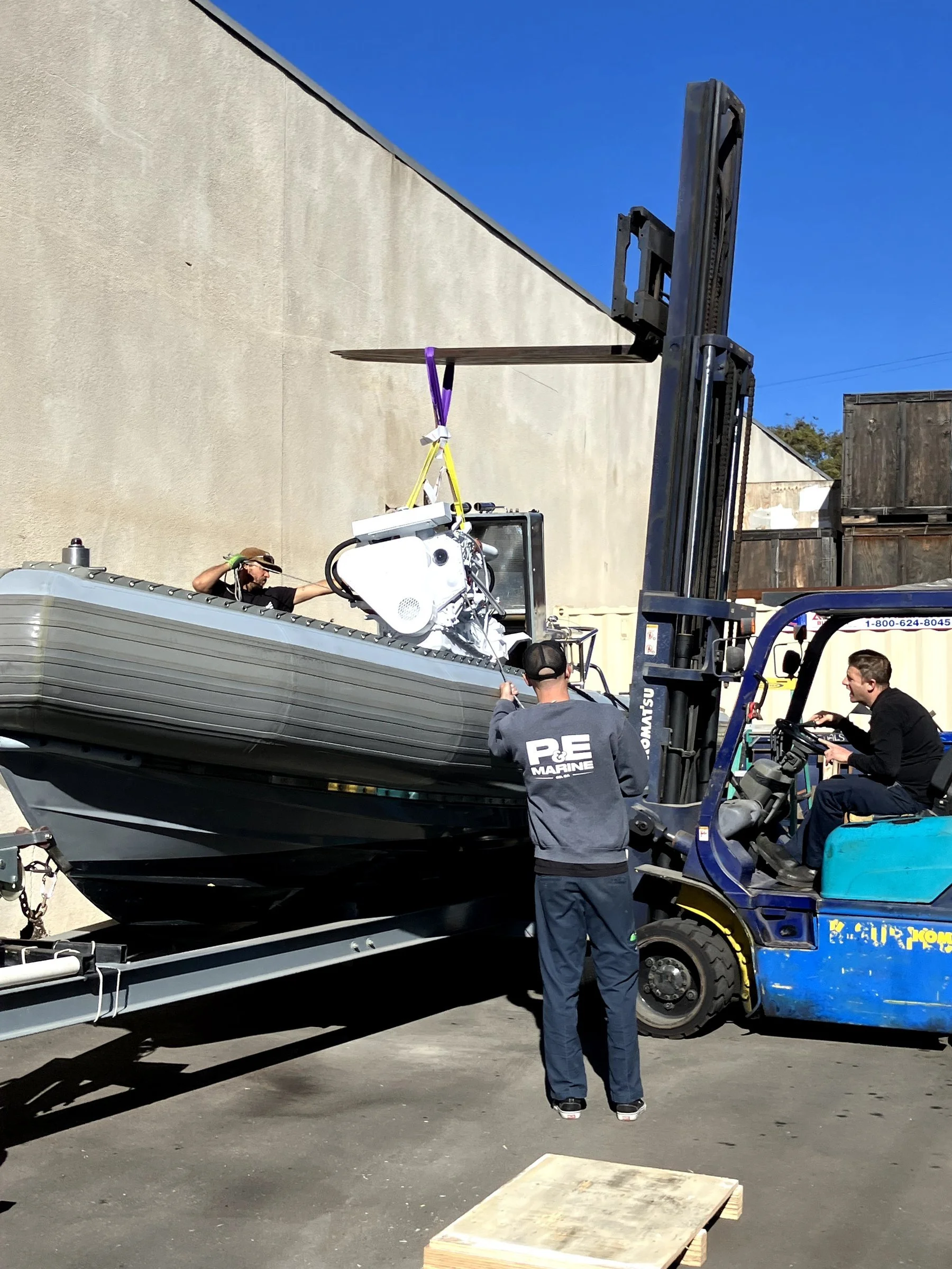 A boat on a trailer being moved by a forklift, with a person in a gray hoodie and another person sitting on the forklift. The scene appears to be outdoors against a beige wall and a blue sky.