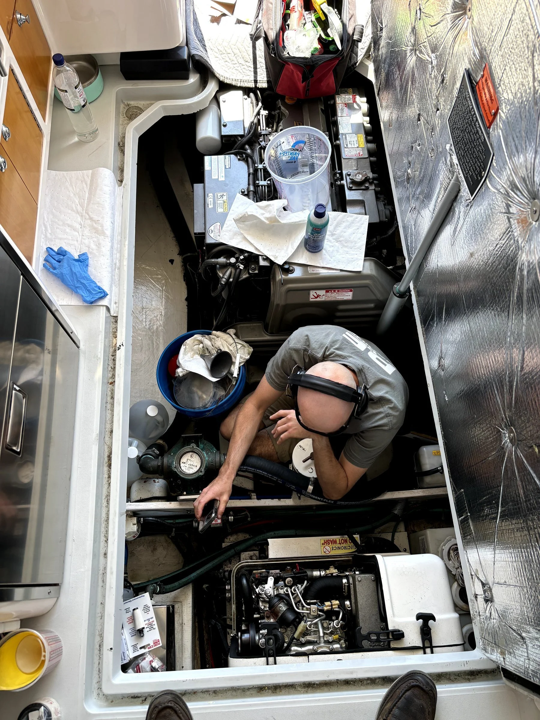 A person wearing a headset working on a mechanical or engine part inside a confined space, possibly an engine compartment or maintenance area, surrounded by tools, supplies, and equipment.