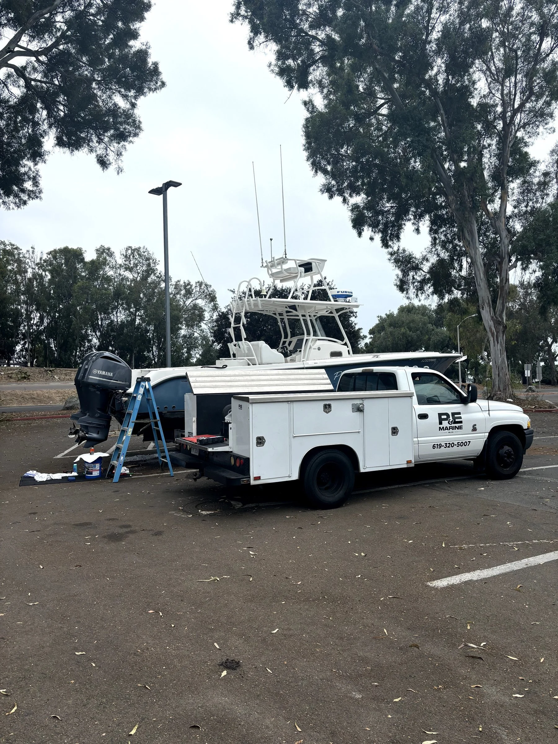 A parking lot with a white boat on a truck, an outboard motor, work supplies, and trees in the background.
