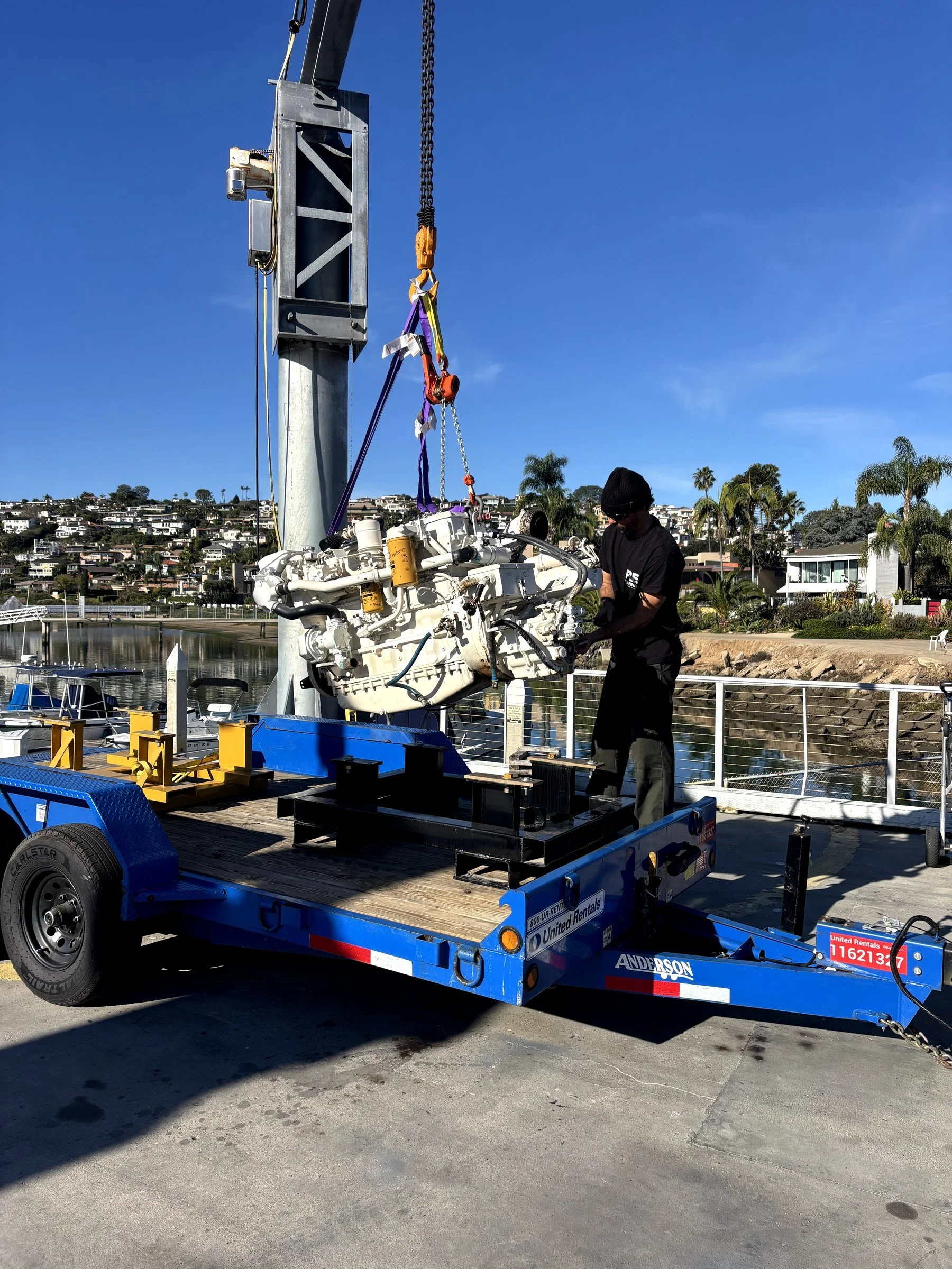 A worker in black clothing and a black beanie manning a boat lift, with a boat engine mounted on a blue trailer, against a backdrop of houses and palm trees under a blue sky.