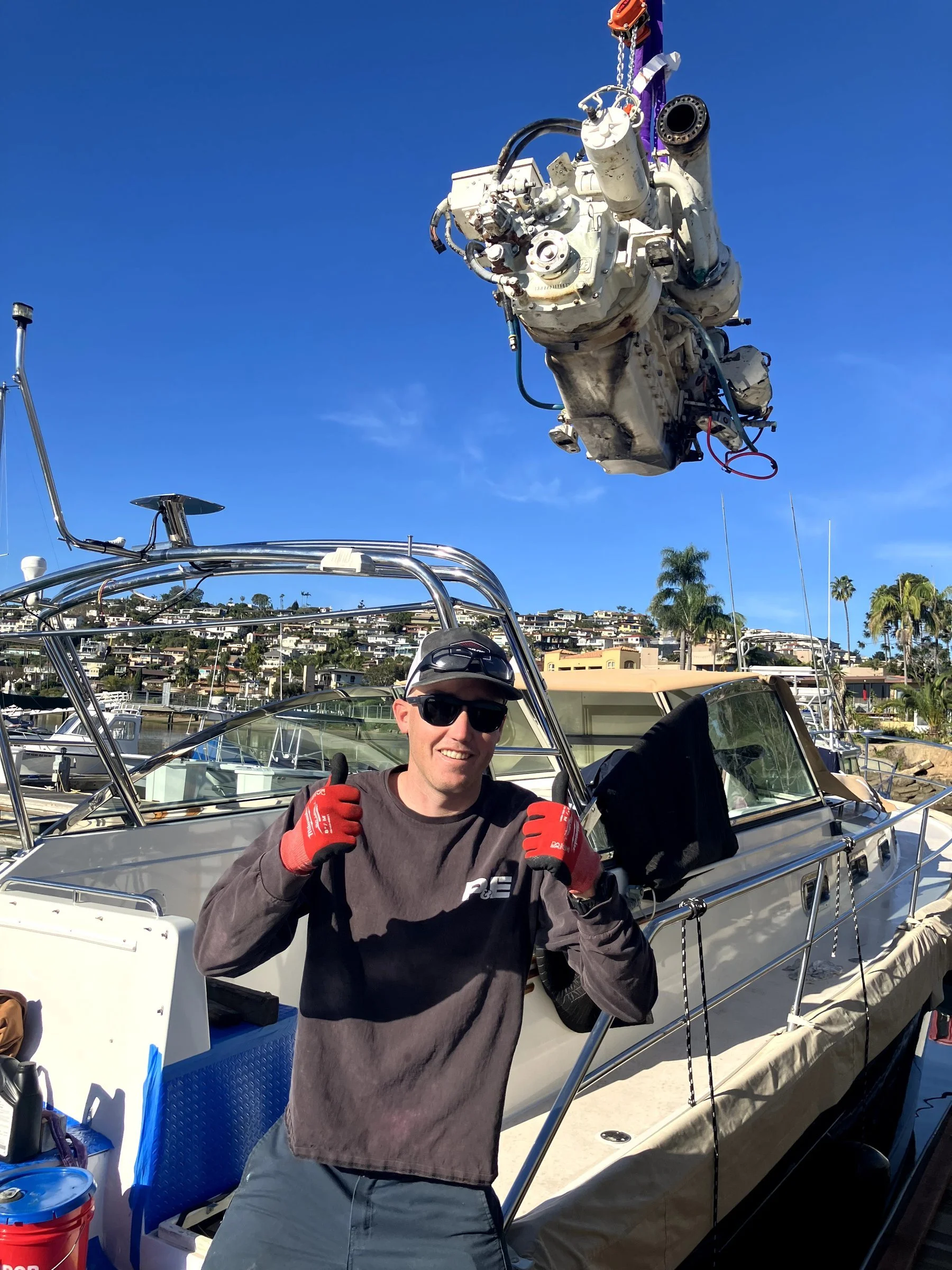A man wearing a dark jacket, red gloves, sunglasses, and a gray cap stands on a boat with a smile, giving two thumbs up. Behind him, there is a large white boat with a visible boat engine suspended overhead, orange safety equipment, and a background 
