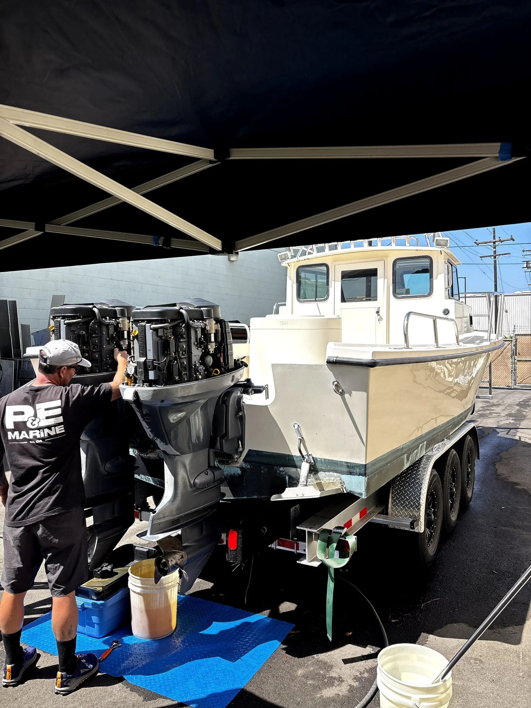 A person working on a boat motor on a trailer under a canopy. The boat is on a trailer, and there are containers, tools, and equipment around.