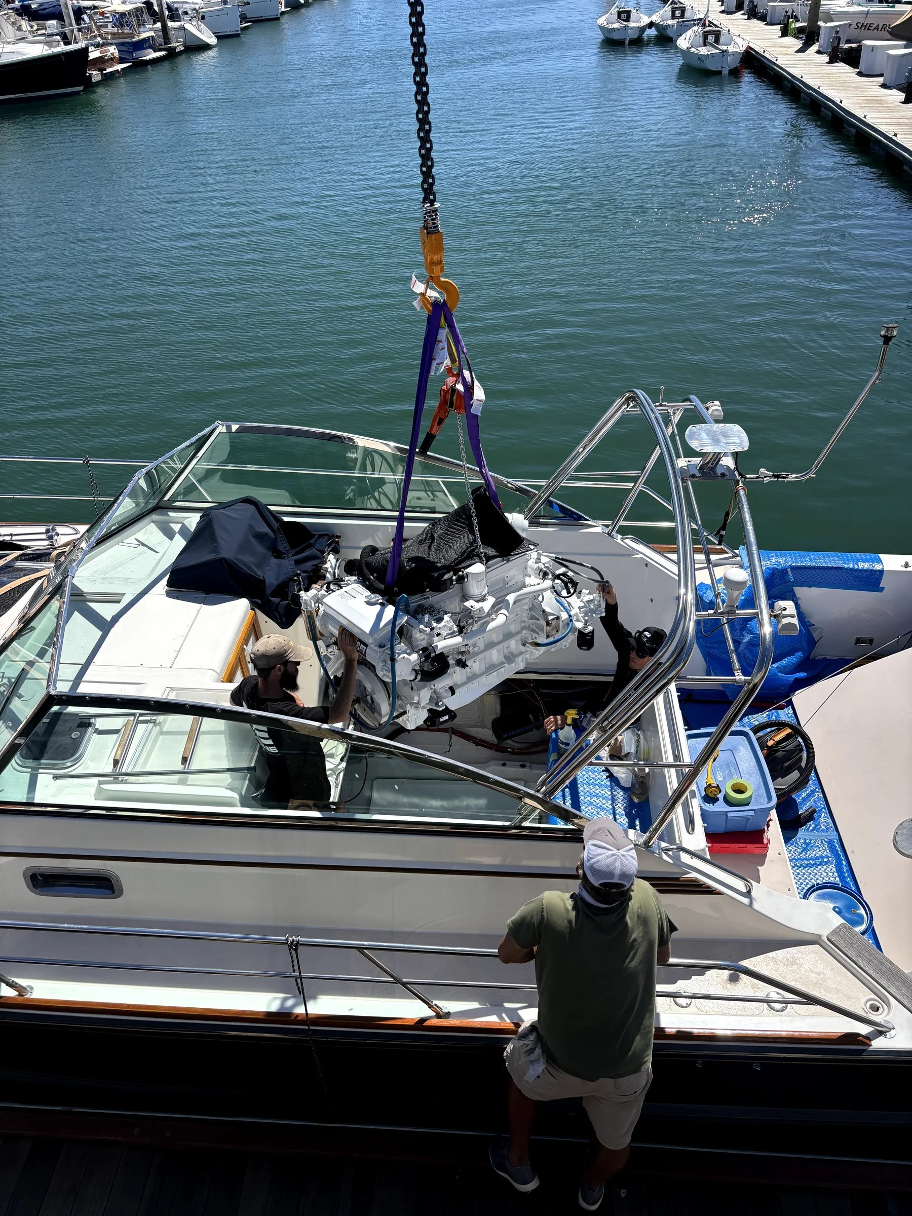 People working on a small boat in a marina, with several other boats docked nearby. One person is standing outside the boat, while others are inside working on the engine.
