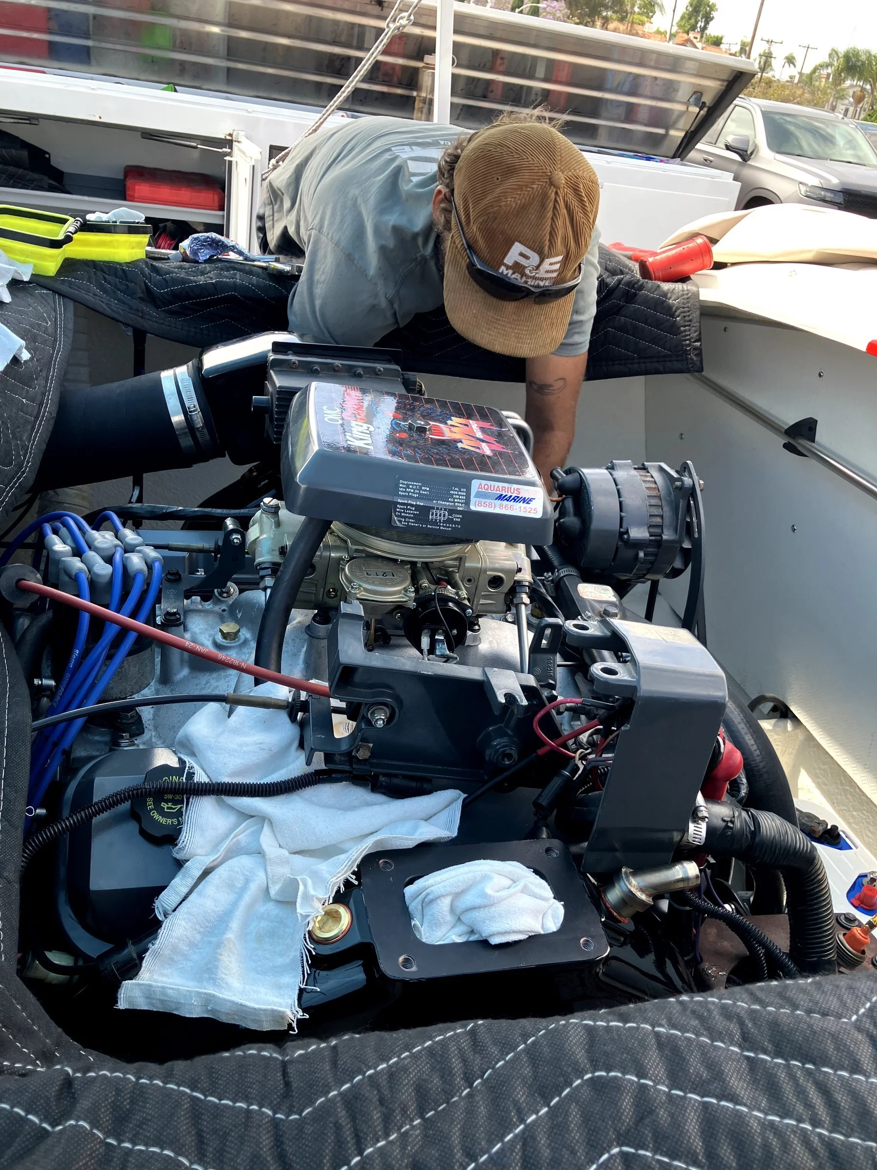 A person wearing a tan cap and glasses working on a car engine, with various tools and car components around, outdoors in a parking lot with palm trees in the background.