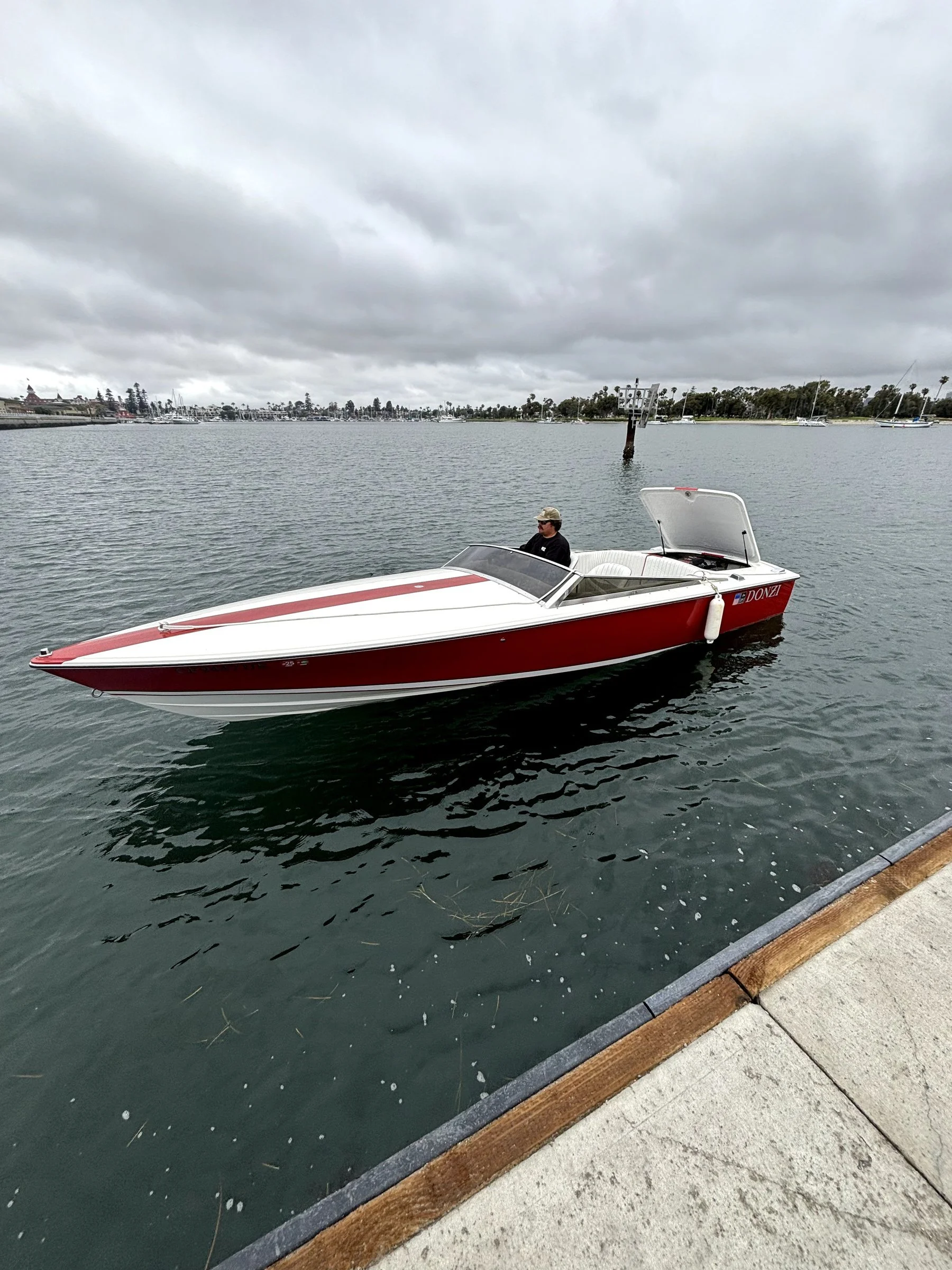 A man in a hat is sitting in a red and white speedboat docked near a pier, with boats and a cloudy sky in the background.