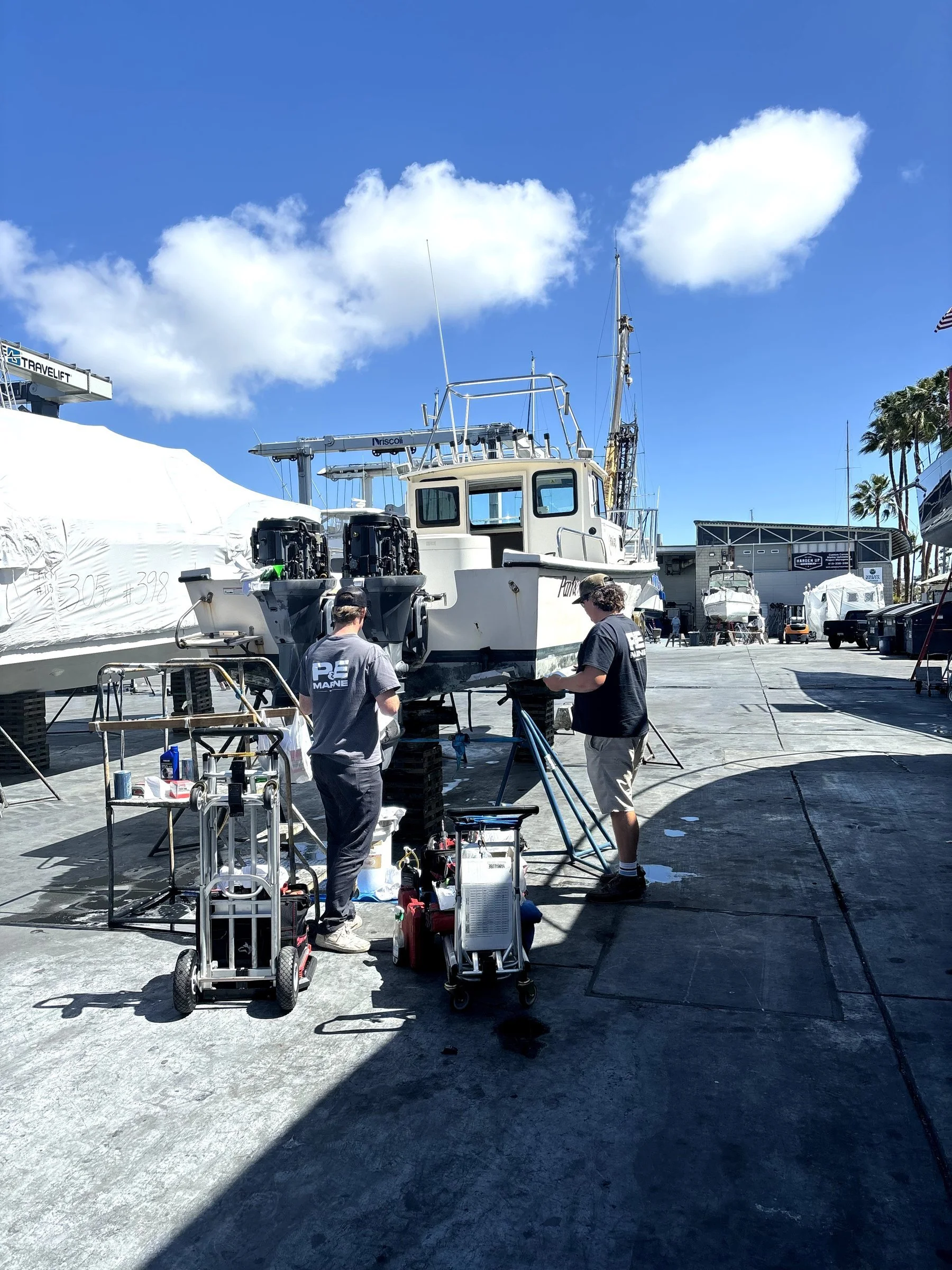 Two men working on a boat in a boatyard. The boat is elevated on supports, with equipment and tools nearby. There are other boats covered or in storage, palm trees, and a building in the background, under a blue sky with scattered clouds.