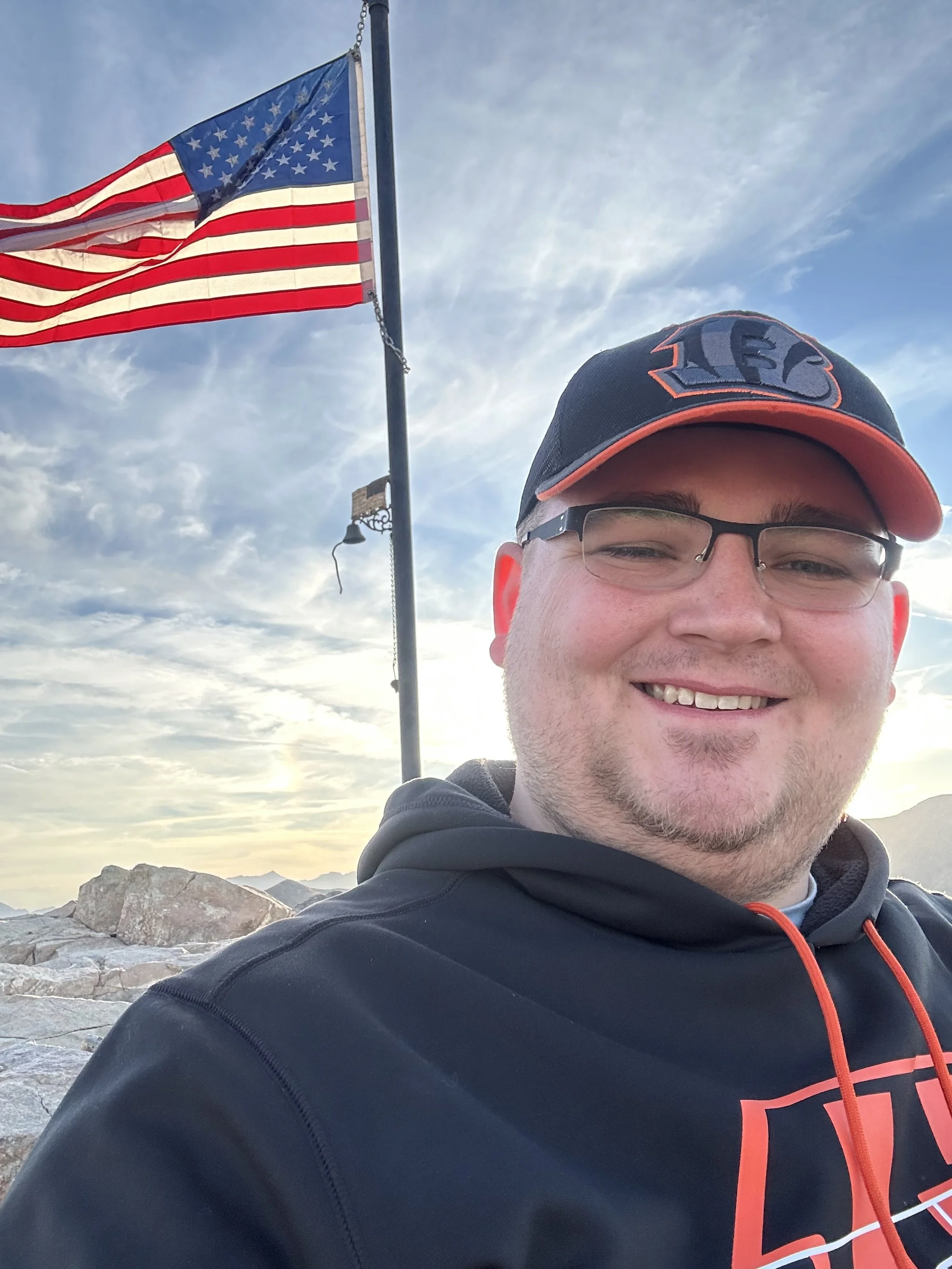 A man taking a selfie outdoors with an American flag flying on a flagpole in the background, a rocky landscape, and mountains in the distance, during sunset or sunrise.