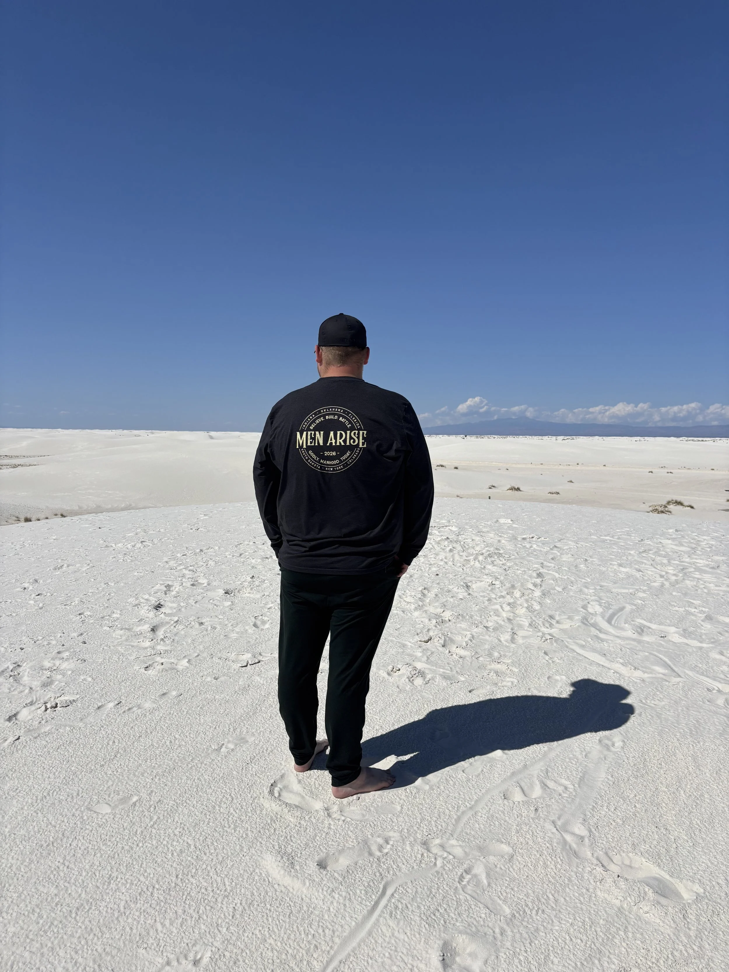 A person standing barefoot on a white sandy landscape under a clear blue sky, wearing a black jacket with 'Men Arise' on the back and a black cap.