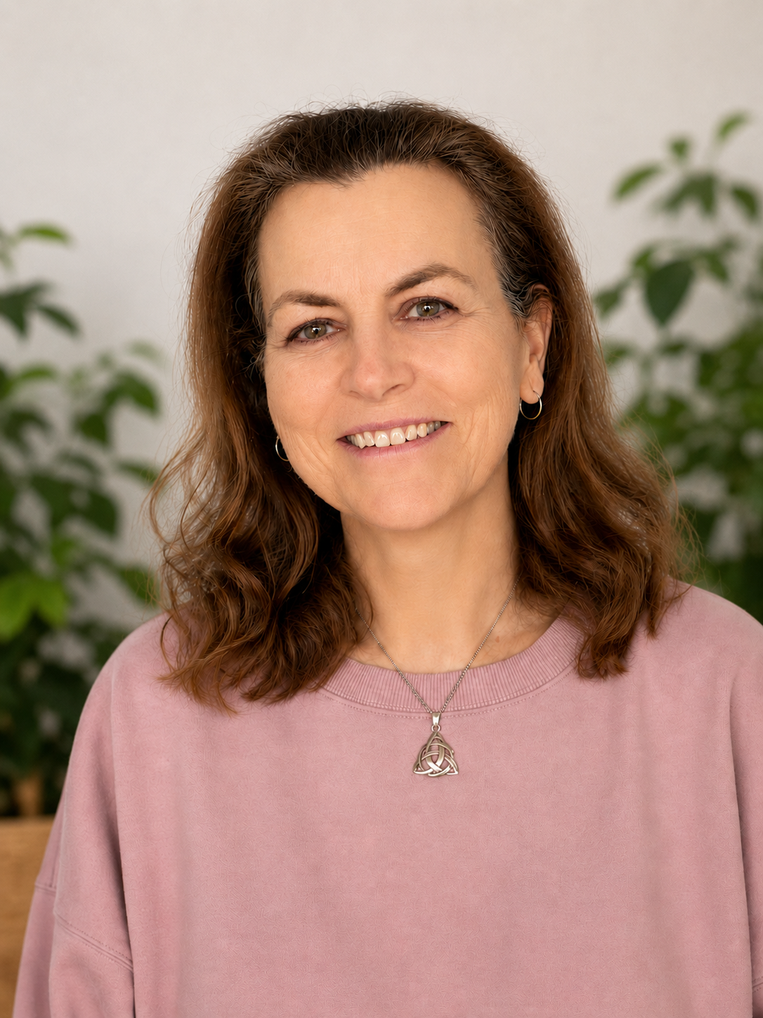 A woman with shoulder-length wavy brown hair, wearing a pink sweatshirt, smiling and looking at the camera, with green plants in the background.
