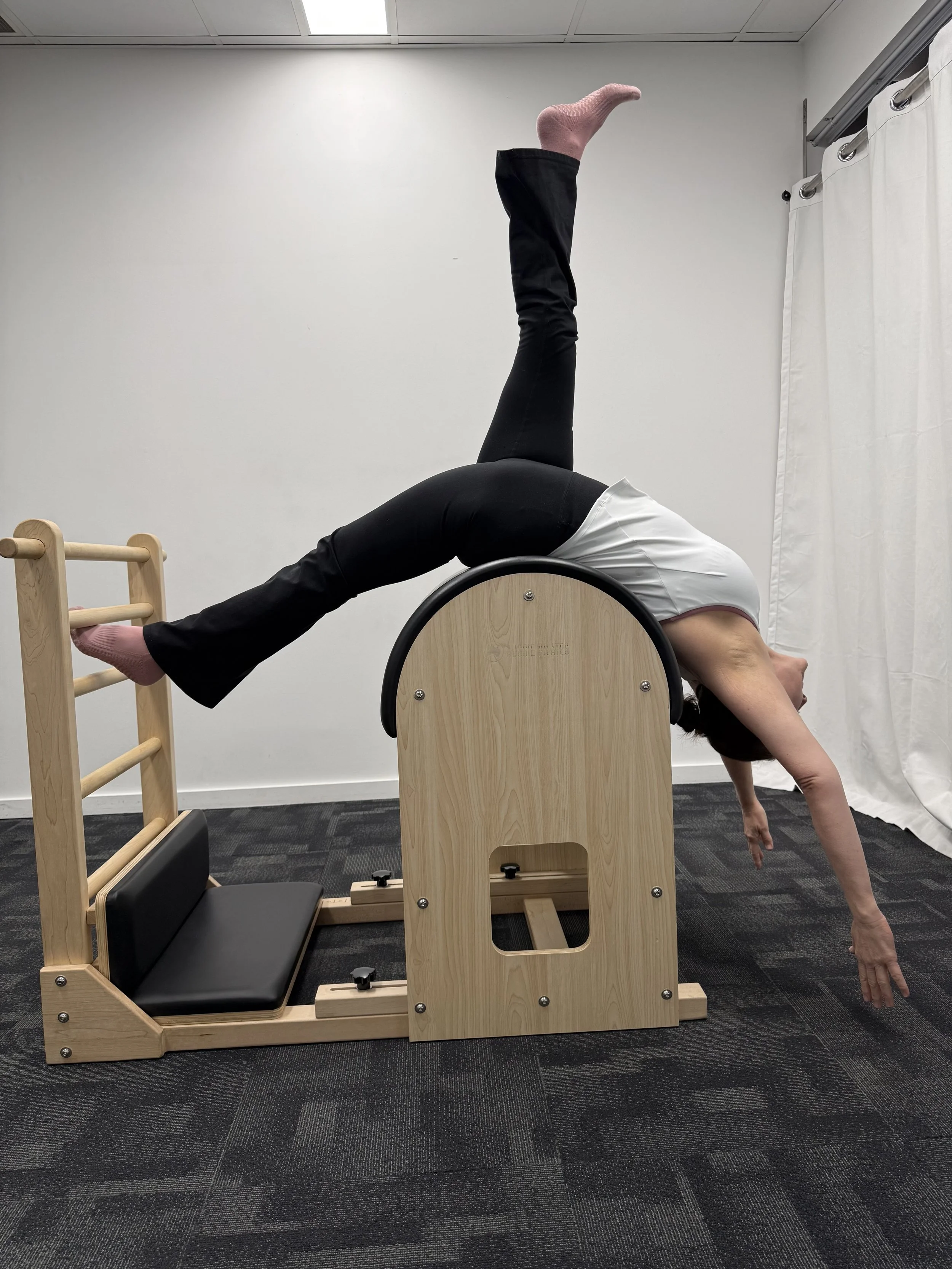 A person performing a yoga pose on a Pilates reformer machine in a studio, with one leg raised straight up and the other arm extended downward, head turned to the side.