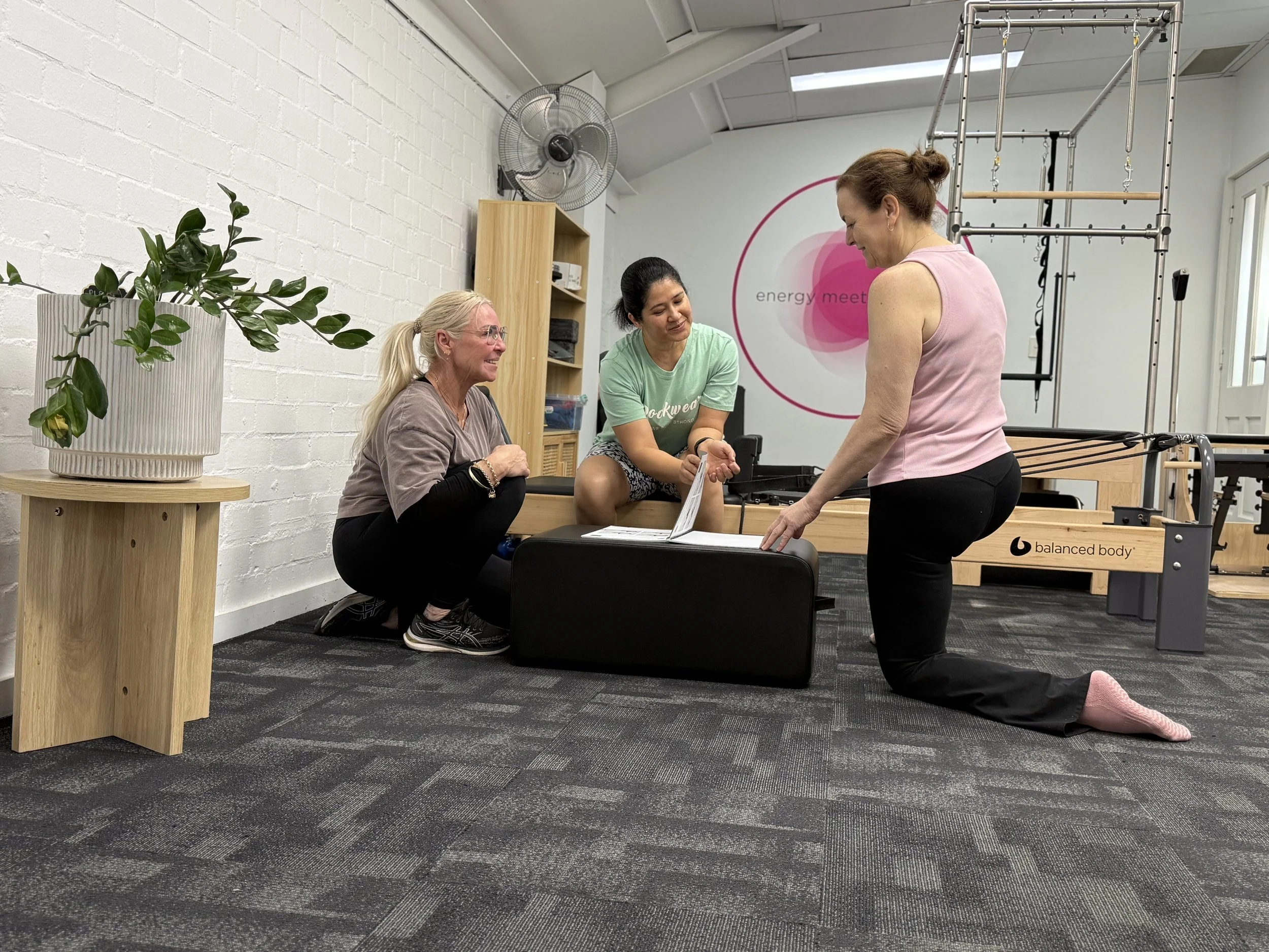 Women in a physical therapy or fitness studio engaging with an instructor around Pilates or rehabilitation equipment, with a potted plant in the foreground and a wall with the branding 'energy meet' in the background.