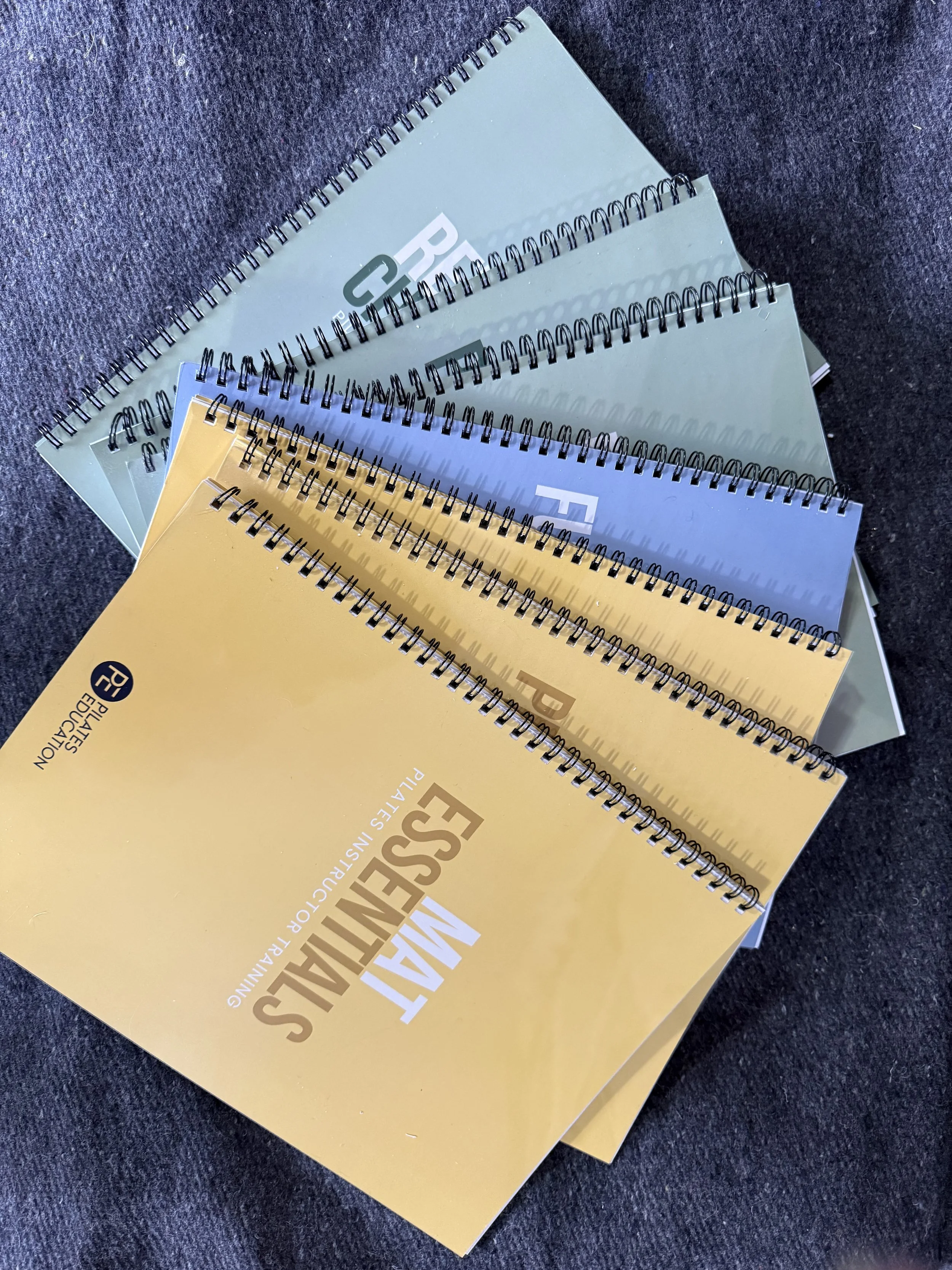 Multiple spiral-bound books or brochures with yellow and light blue covers, lying on a dark fabric surface.