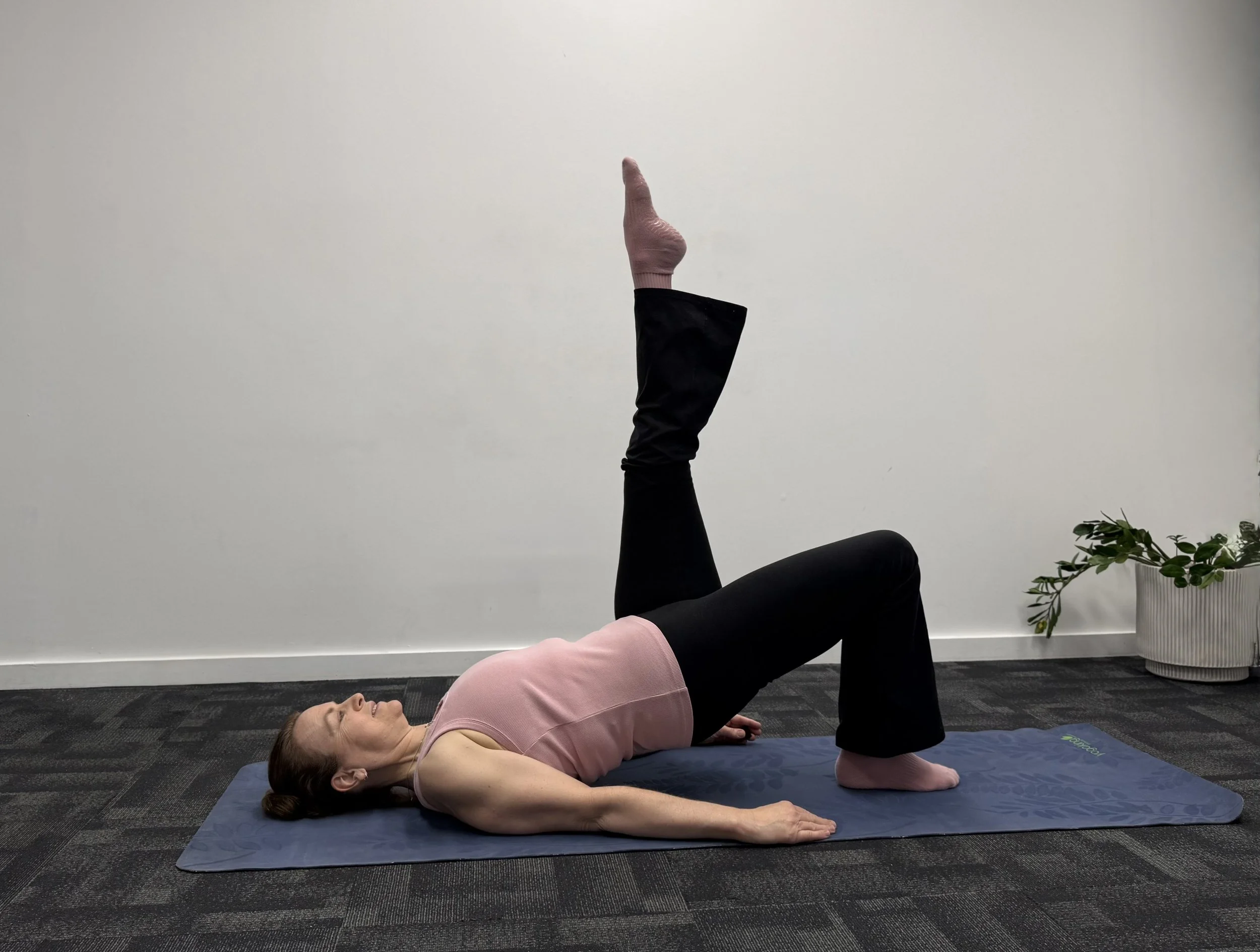 A woman lying on her back on a yoga mat with her right leg raised straight up, supported by her raised hand, while her left leg is bent at the knee and her arms are relaxed on the floor.